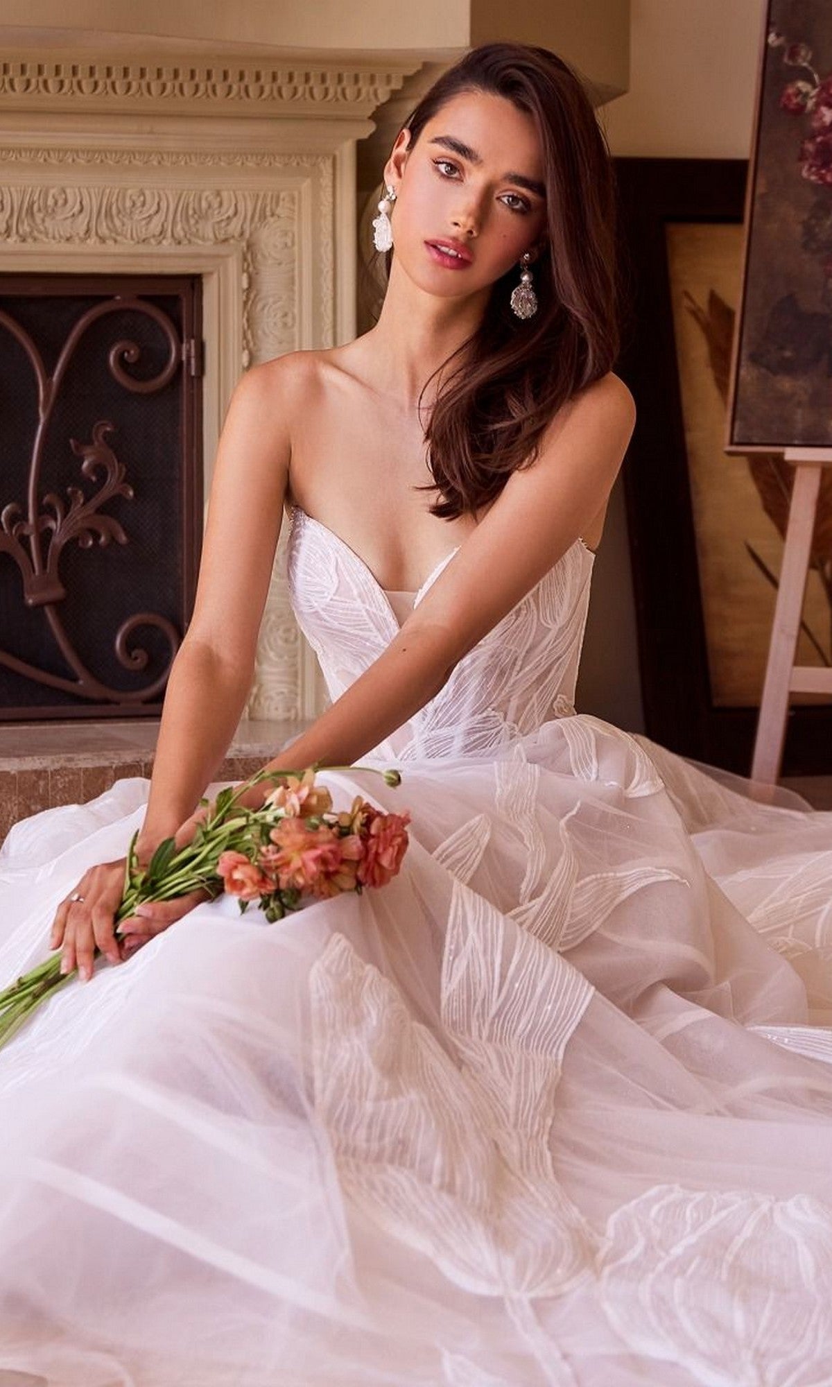 A woman wearing the Andrea and Leo WL043 long wedding dress with a tulle A-line skirt sits indoors holding a bouquet, with a fireplace and artwork in the background.