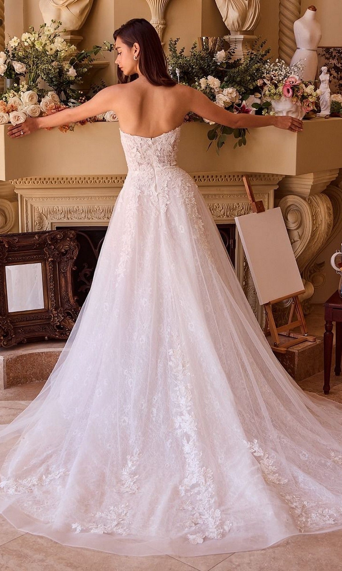 A woman wearing the Andrea and Leo WL052 long wedding dress, a white strapless gown with delicate floral lace applique, stands with arms outstretched in front of a flower-adorned fireplace mantel.