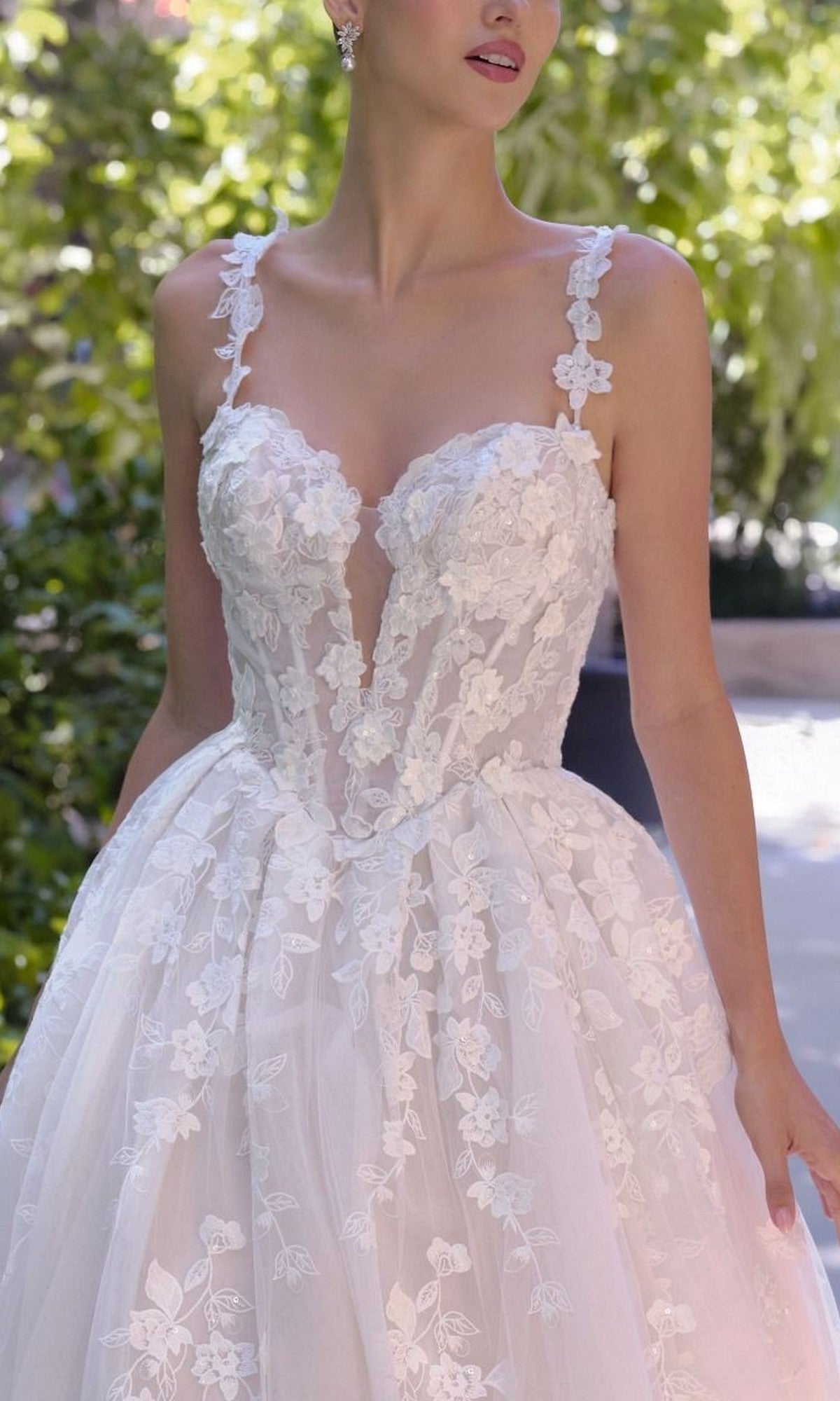 A woman stands outdoors amid greenery wearing the Andrea and Leo WL058 long wedding dress, featuring white floral lace, a deep neckline, spaghetti straps, and a full A-line skirt.
