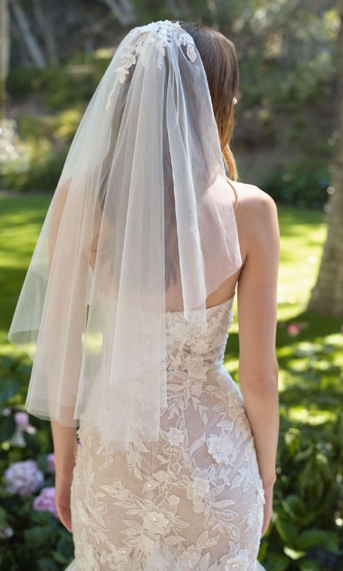 Bride wearing a lace wedding dress and veil in a garden setting, facing away from the camera to show the back.