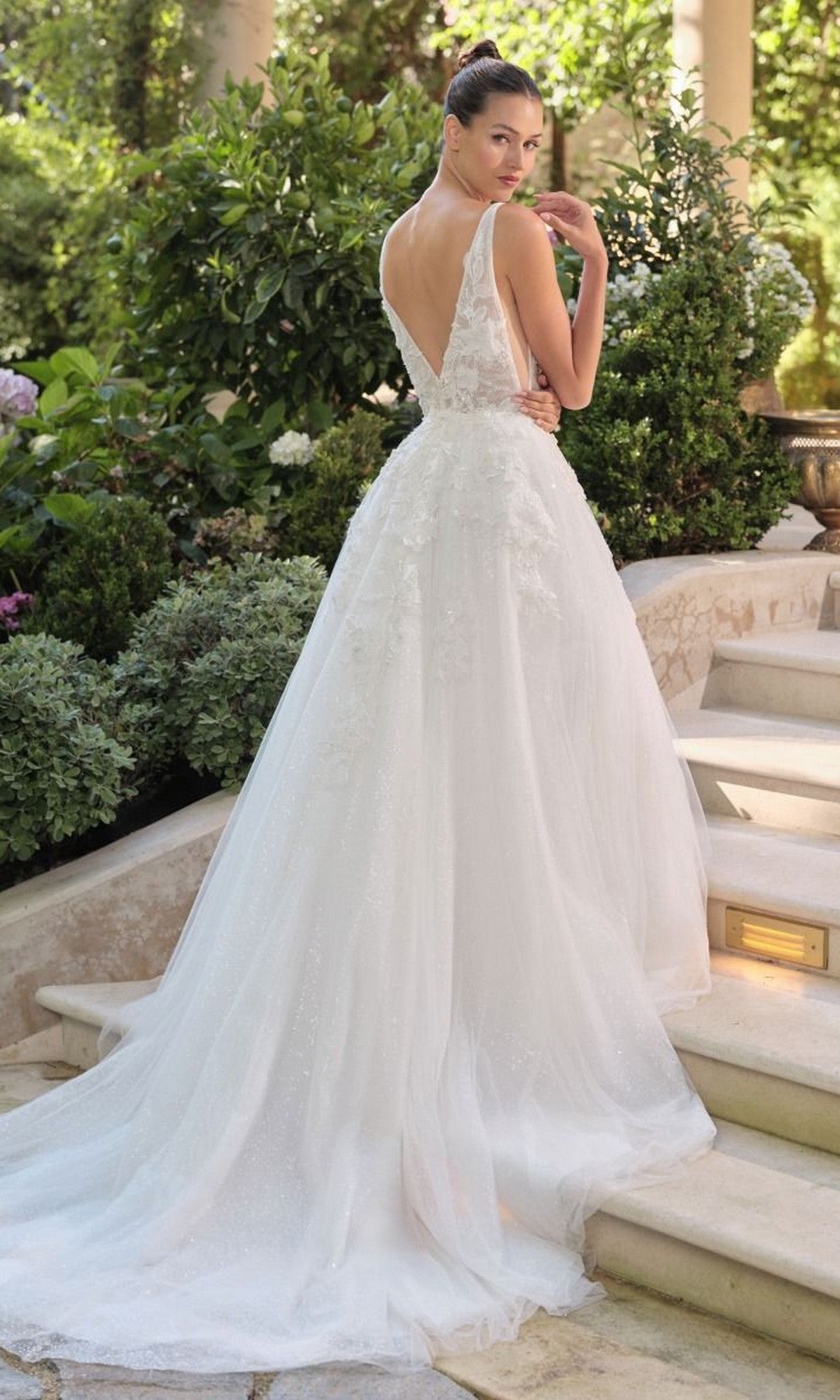 A woman wearing the Andrea and Leo WL064 long wedding dress, featuring delicate lace appliques and a flowing tulle skirt, stands on outdoor stone steps surrounded by lush greenery.