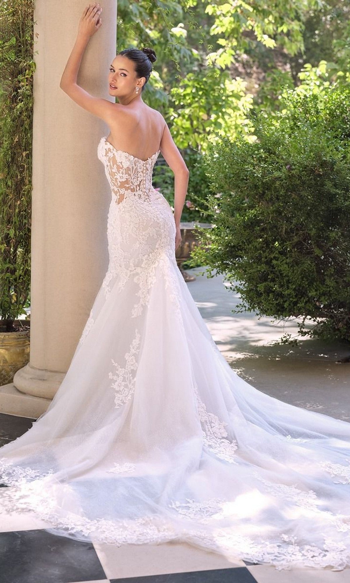 A woman in the Andrea and Leo WL065 Long Wedding Dress, featuring a strapless lace mermaid silhouette and long train, stands outdoors touching a column and looking over her shoulder.