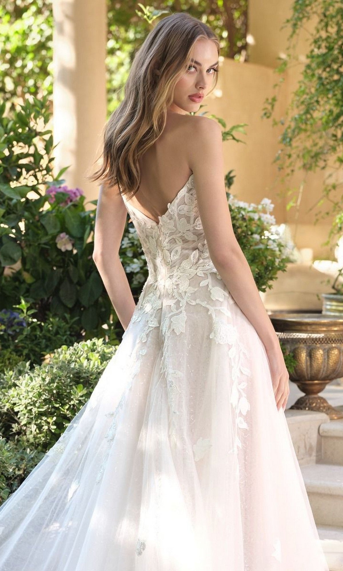 A woman with long, wavy hair stands outdoors wearing the Andrea and Leo WL070 Long Wedding Dress, which features 3D floral lace appliques and a low back. Greenery and flowers can be seen in the background.
