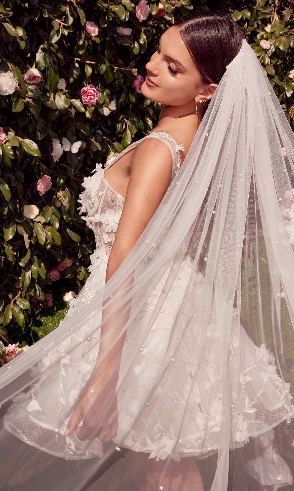 A woman in the Andrea and Leo WL081 short white dress and a long veil stands before lush green leaves and pink flowers, looking down with a soft expression.