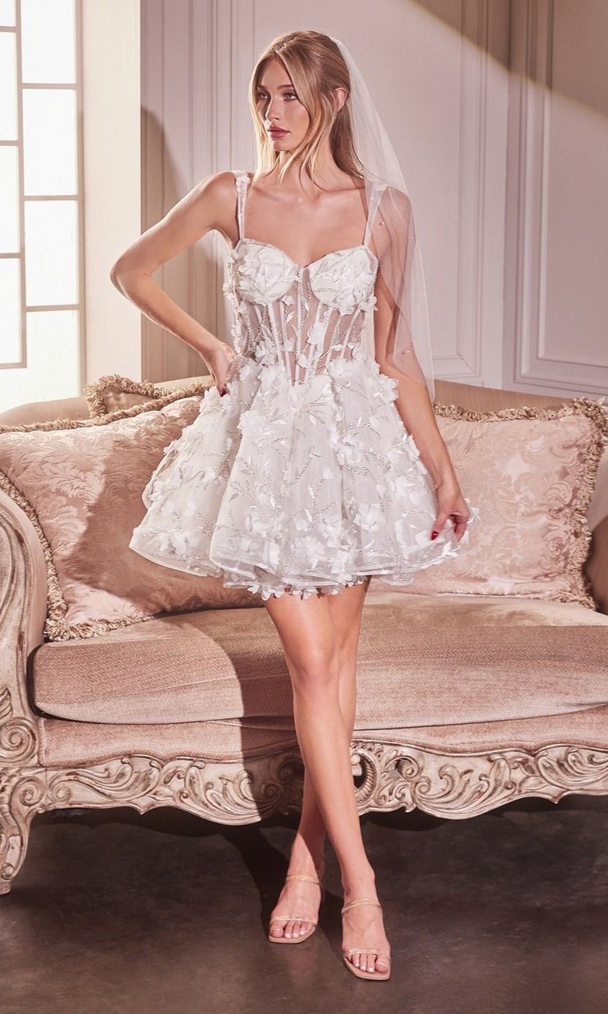 A woman wearing the Andrea and Leo WL081 short white dress with 3D floral lace and a veil stands before an ornate sofa in a softly lit room.