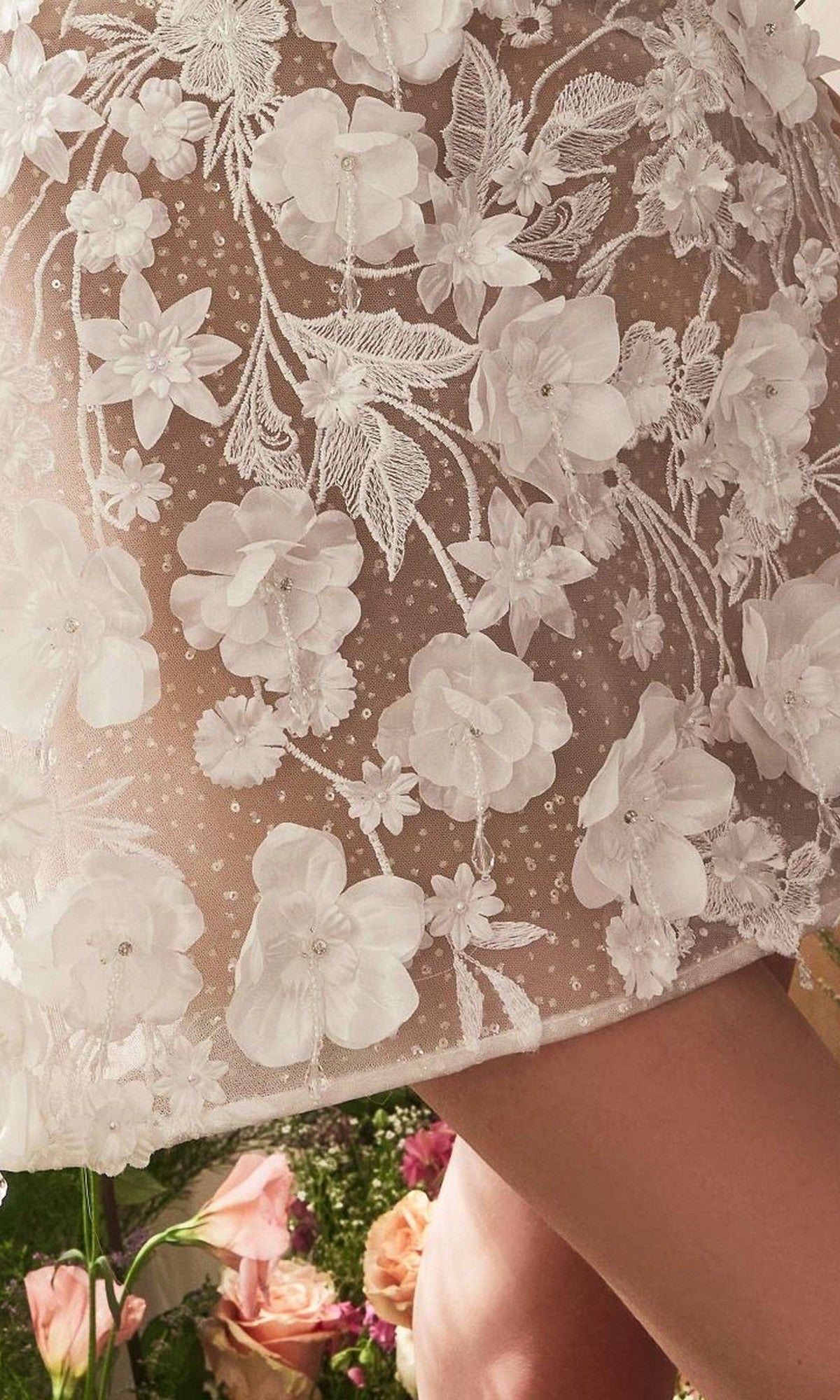 Close-up of the Andrea and Leo WL085 short white dress, featuring detailed 3D floral lace embroidery and appliqué. A bare leg is visible beside assorted flowers in the background.