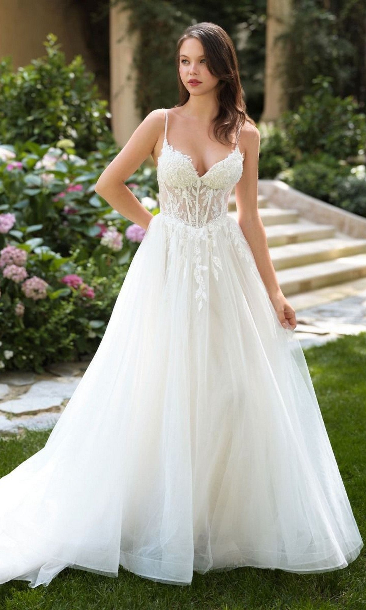 A woman stands outdoors in the Andrea and Leo WL087 long wedding dress, featuring an A-line silhouette with lace appliques, a sheer bodice, and a flowing tulle skirt. Garden flowers and stone steps can be seen in the background.
