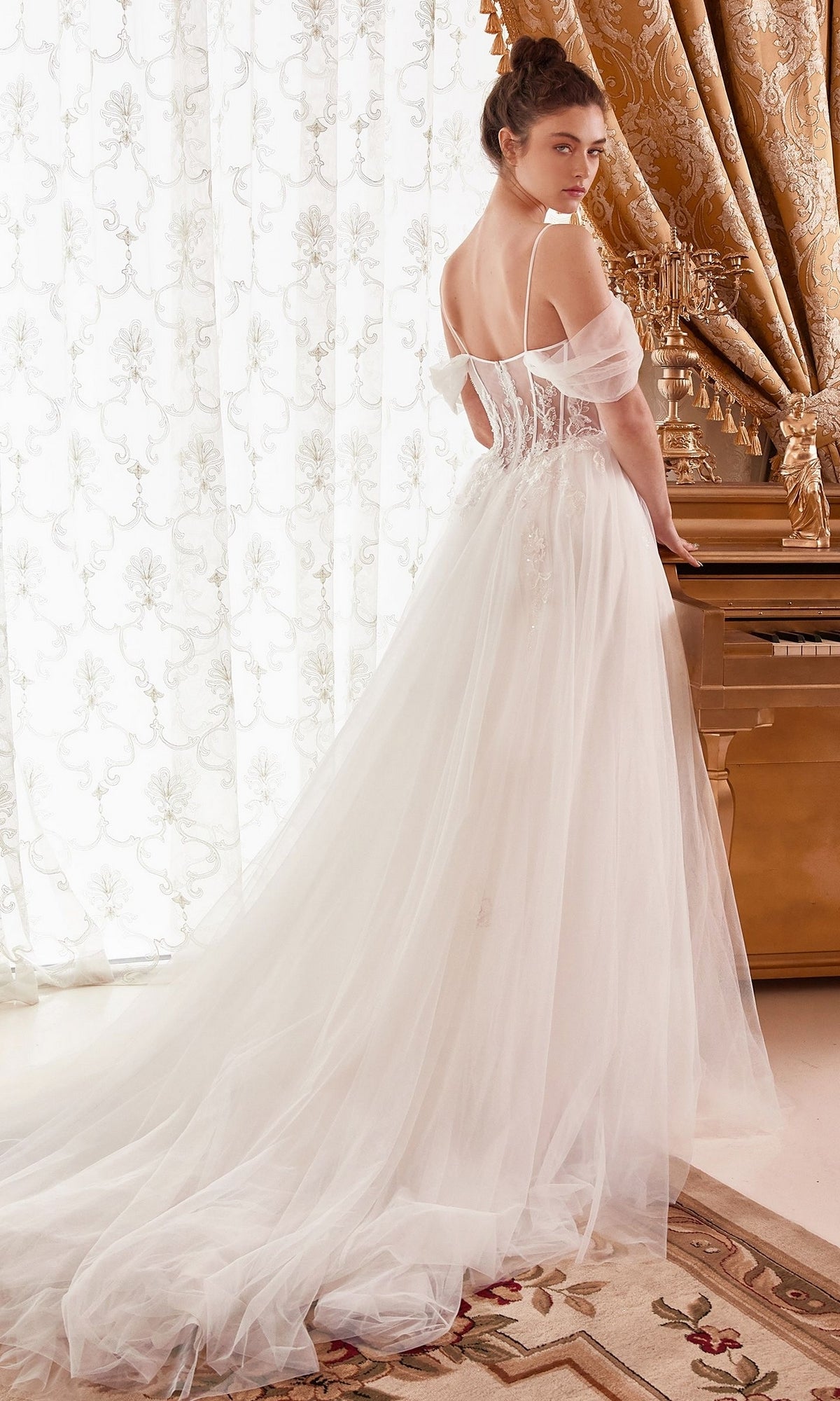 Wearing the Off-White Sheer-Bodice Long Ball Gown WN307, a woman stands by a gold piano and ornate curtains, gracefully looking over her shoulder.