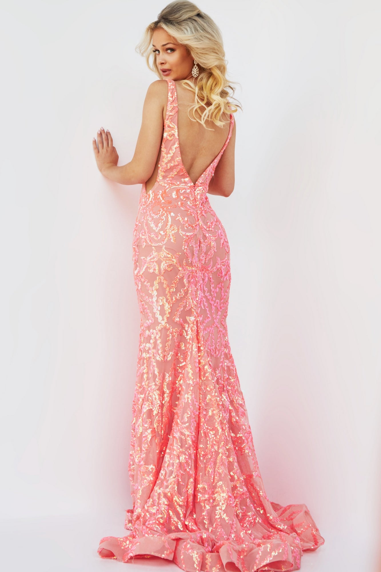 A woman wearing a floor-length, sleeveless Jovani Sequin Prom Dress with a plunging neckline and low back in peach, standing against a white background.