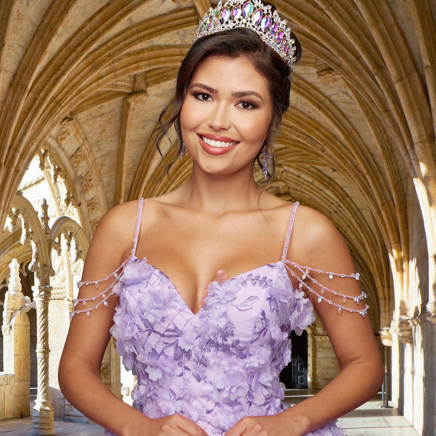 A woman wearing the Quinceañera Sample Dress QF105, a lavender cold-shoulder gown with floral details and a jeweled crown, smiles at the camera while standing in a stone archway.