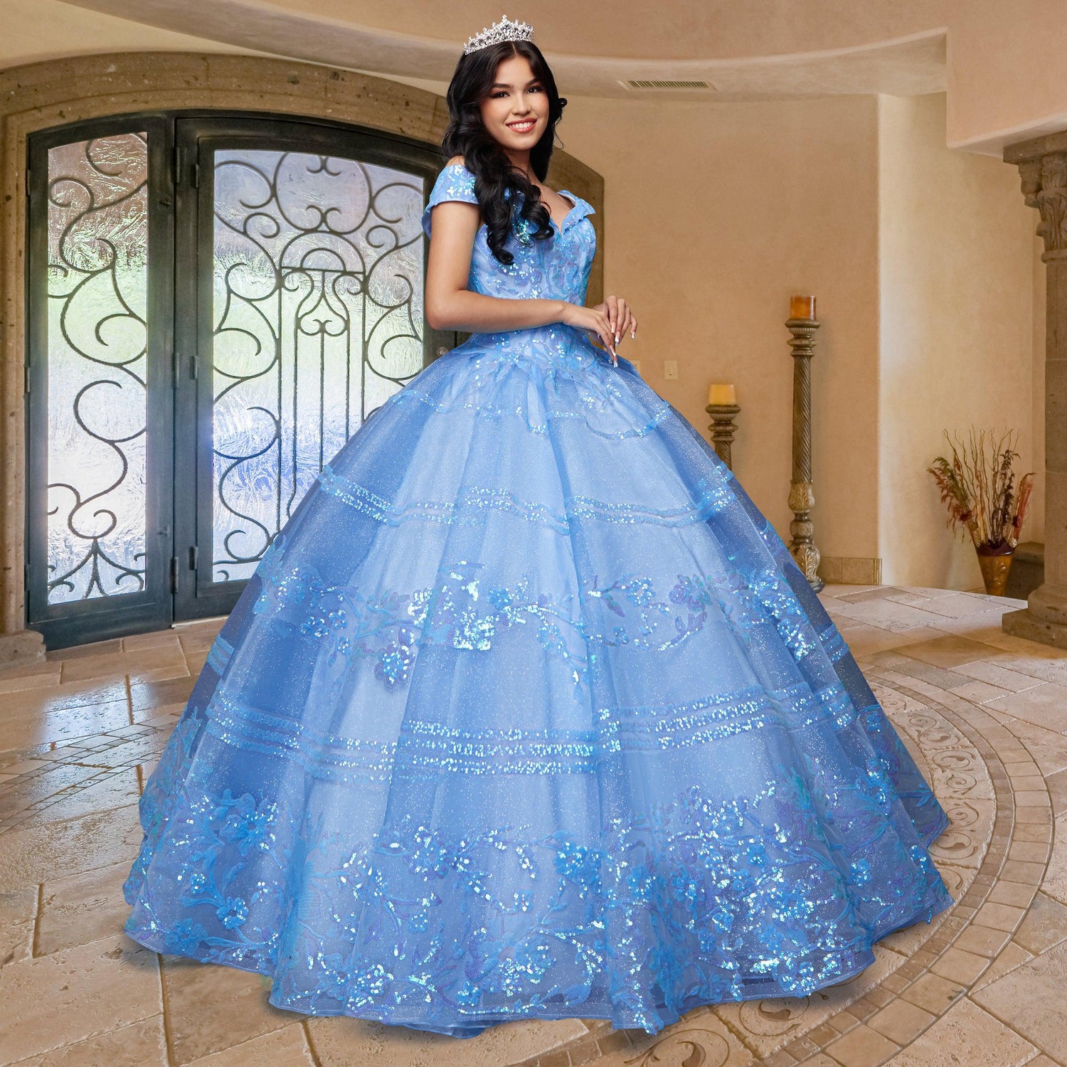 A young woman stands indoors, wearing the sparkling blue Quinceañera Sample Dress QF104 and a tiara, with her hands clasped in front of her.