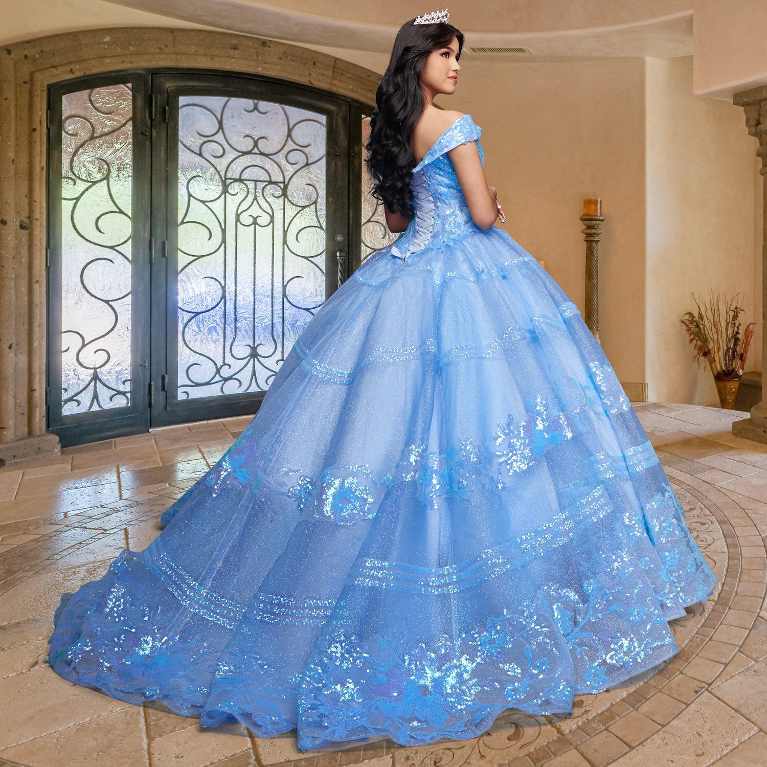A young woman wearing the Quinceañera Sample Dress QF104, a blue off-the-shoulder gown, and a tiara stands indoors on a tiled floor, facing decorative double doors with her back to the camera.
