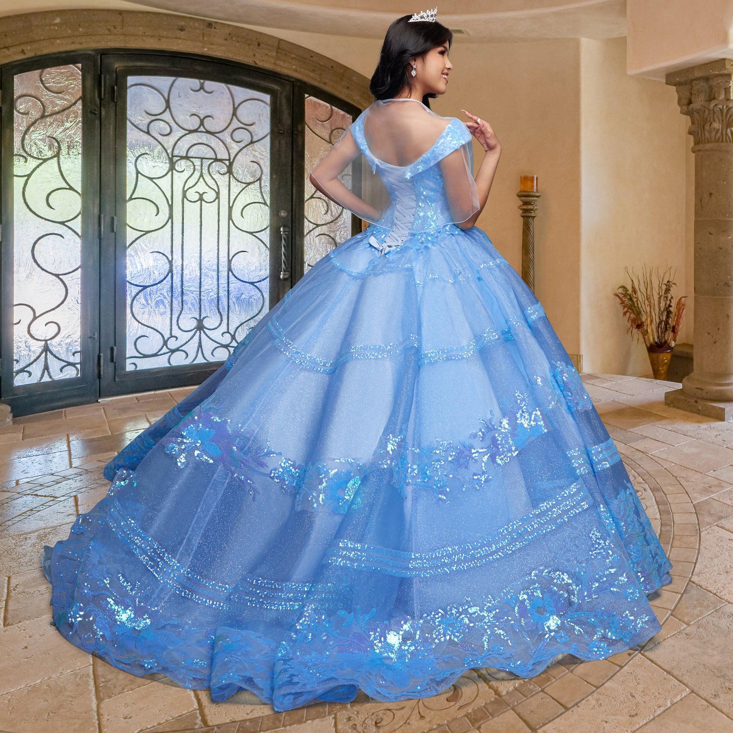 A young woman wears the Quinceañera Sample Dress QF104 and a tiara, standing indoors on a tiled floor facing ornate glass doors with her back to the camera.