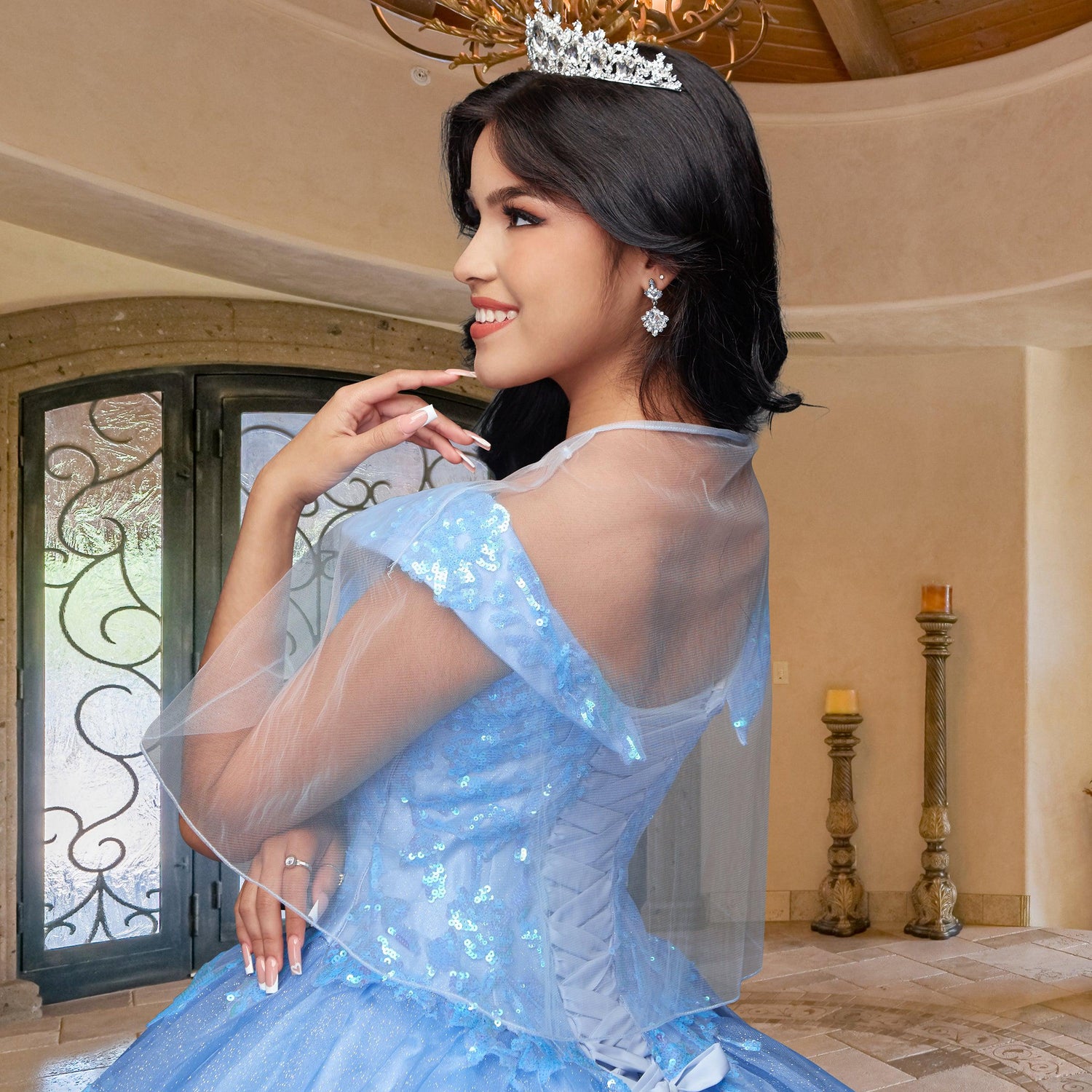 A woman wearing the Quinceañera Sample Dress QF104 and a tiara smiles indoors near a decorative door and candle holders.