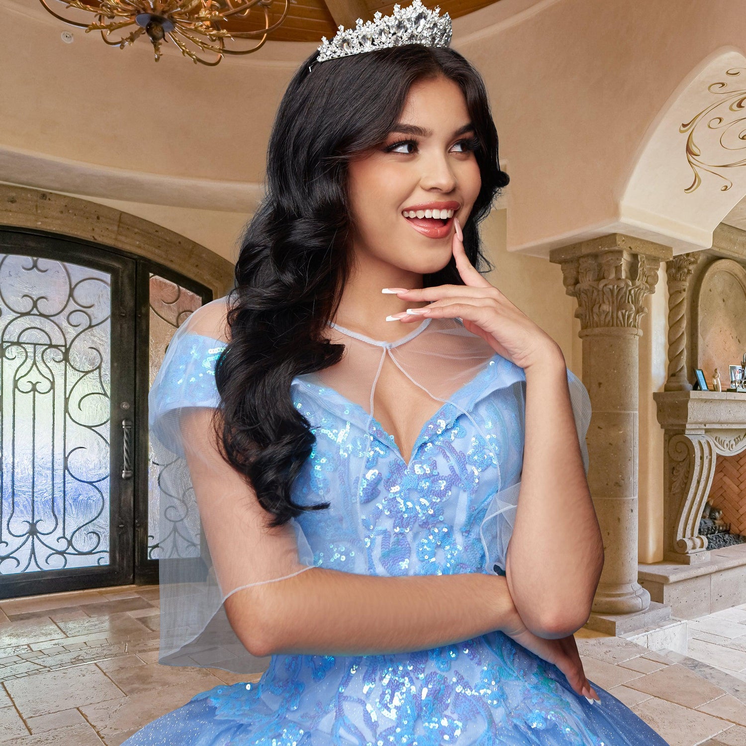 A young woman wears the Quinceañera Sample Dress QF104, a blue, sparkly off-shoulder gown with a tiara, posing indoors with a smile in an elegant room featuring arched doorways and ornate decor.
