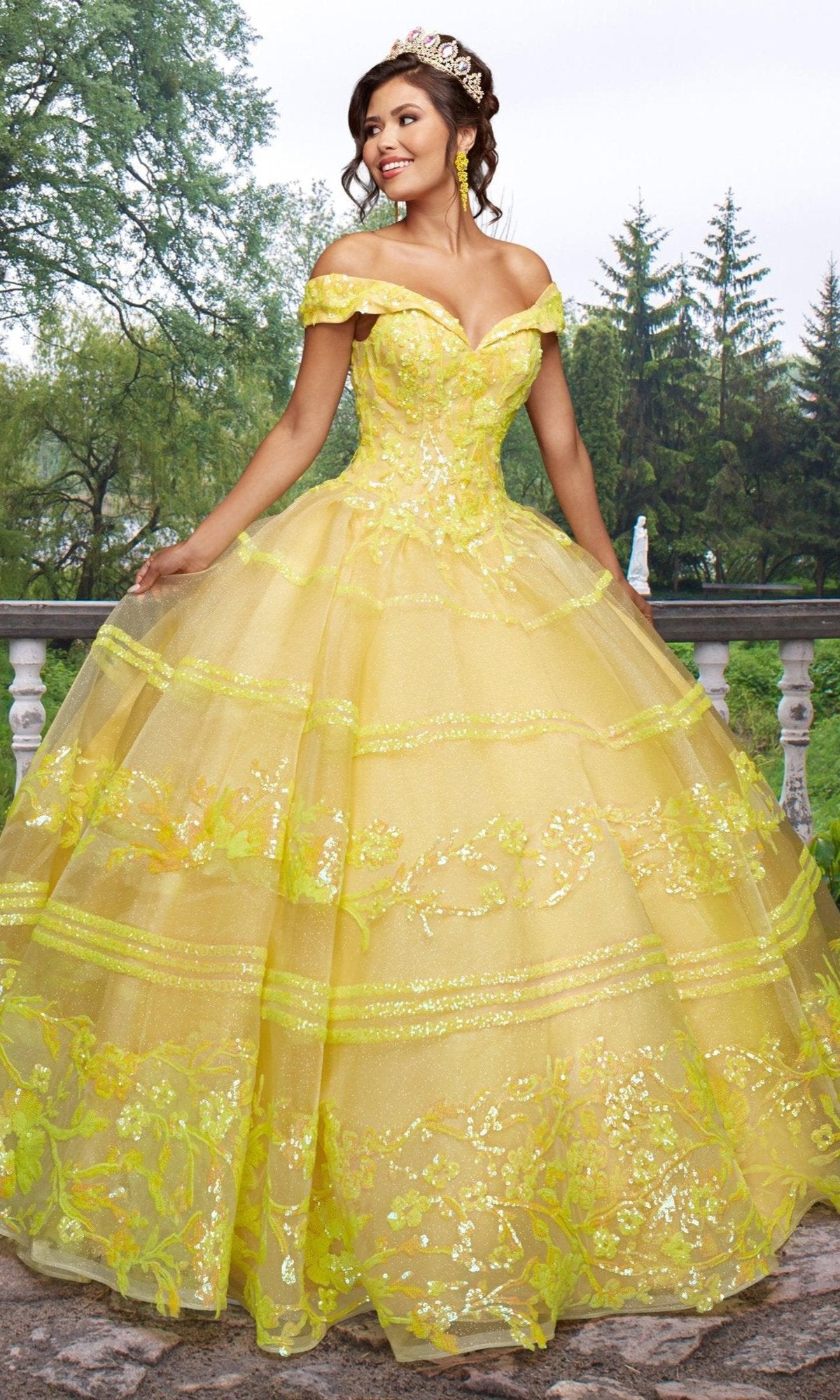 A young woman stands outdoors wearing the Quinceañera Sample Dress QF104, an off-the-shoulder gown with sequin details, complemented by a sparkling tiara.
