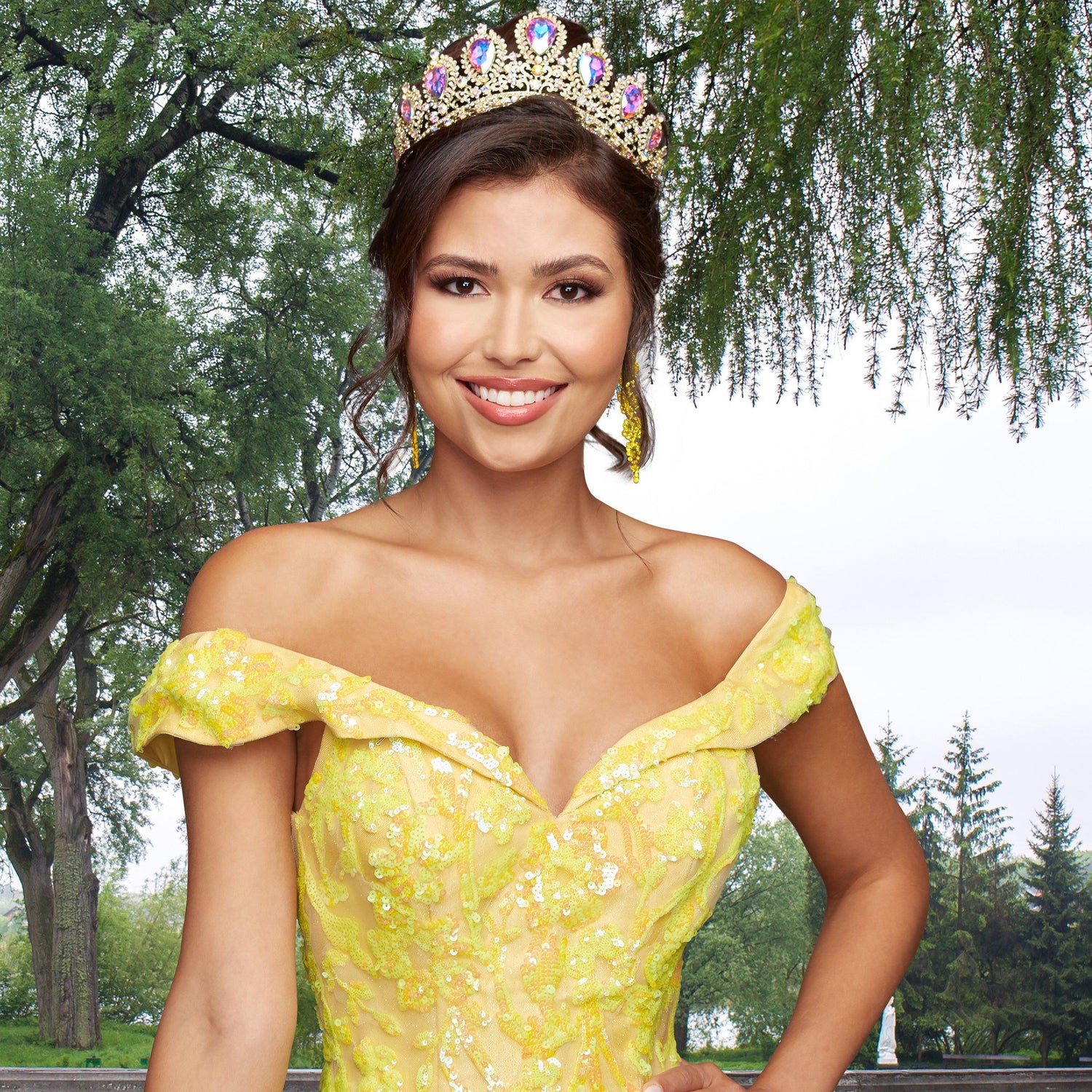 A woman wears the Quinceañera Sample Dress QF104 and a jeweled crown, smiling radiantly outdoors with lush trees in the background.