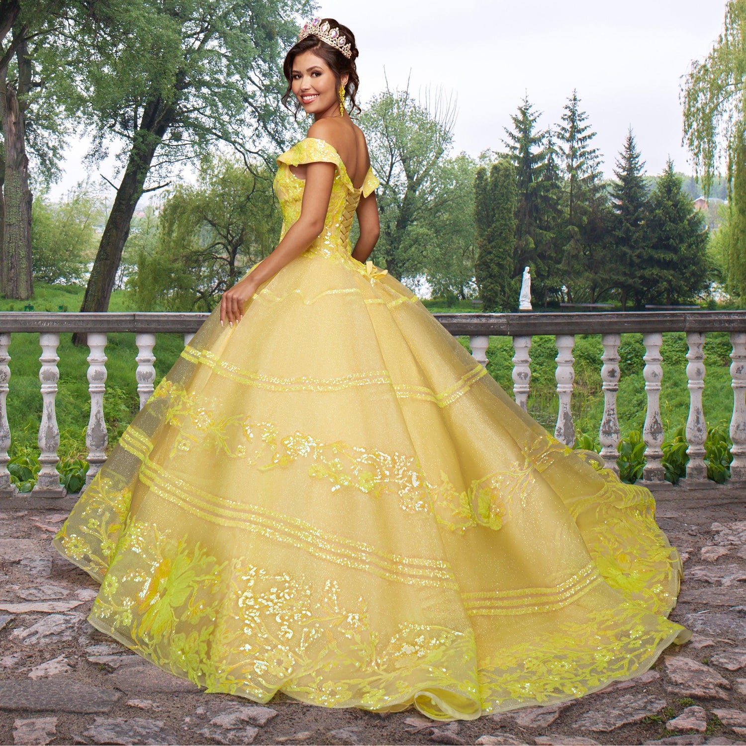 A woman wearing the Quinceañera Sample Dress QF104 with a tiara stands outdoors on a stone patio, surrounded by trees and greenery.