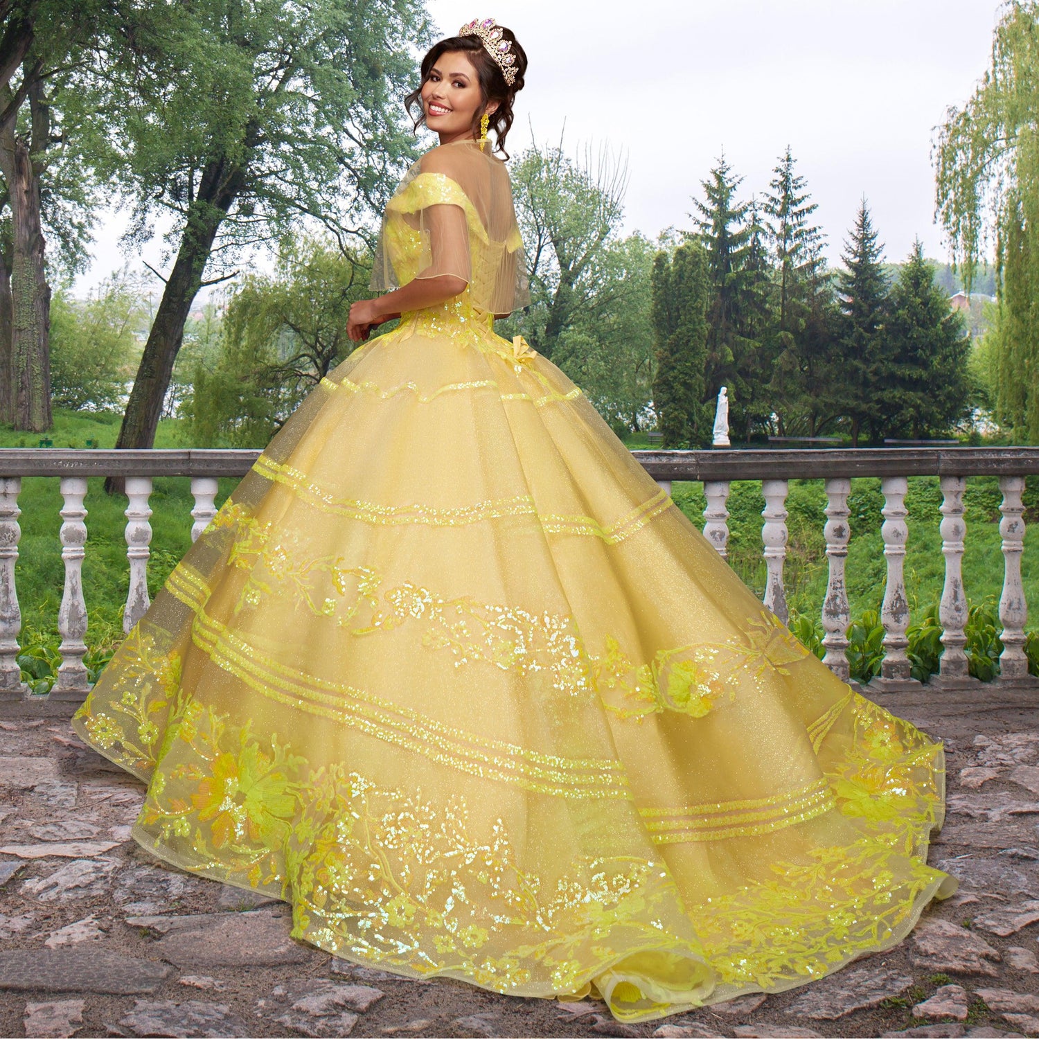 A woman wearing the Quinceañera Sample Dress QF104 and a tiara stands outdoors on a stone terrace, surrounded by trees and greenery.