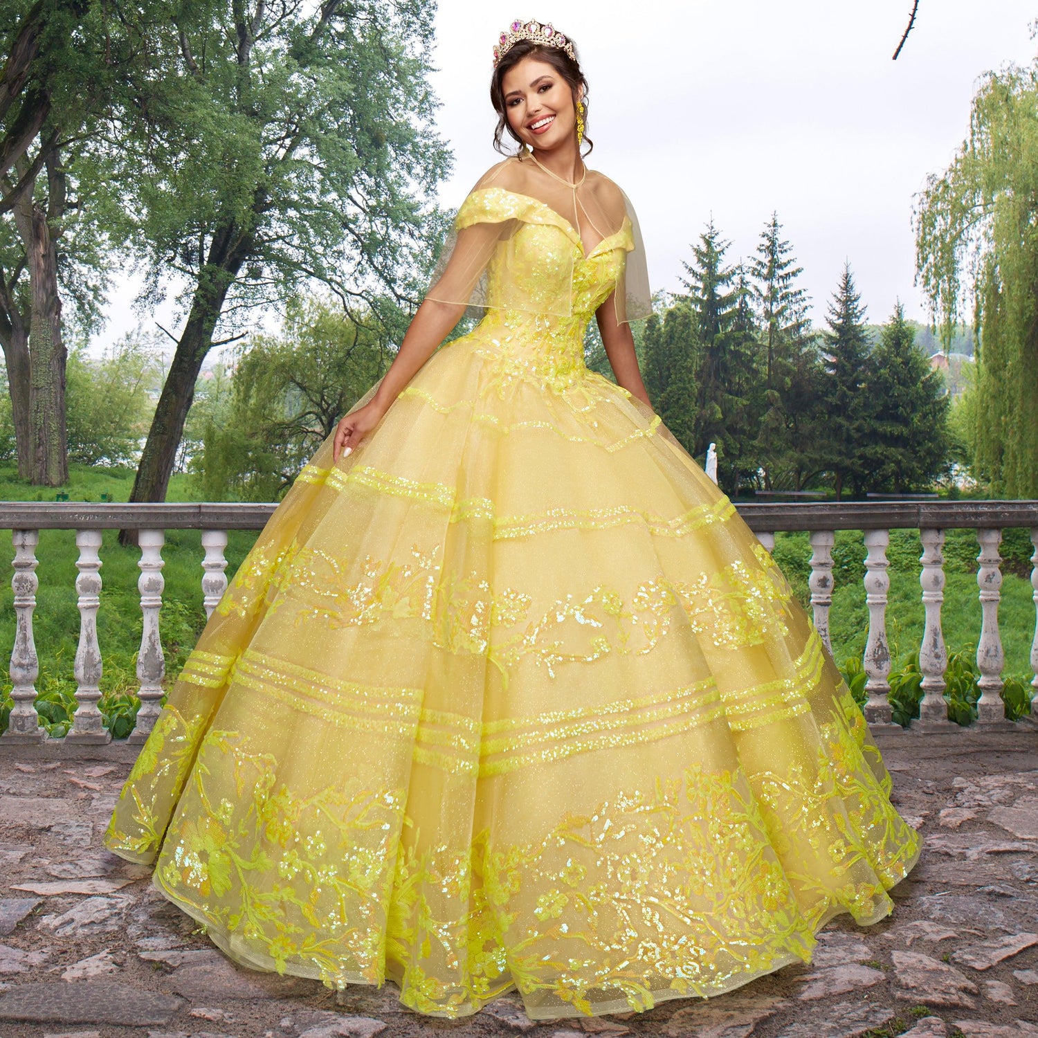 Wearing the Quinceañera Sample Dress QF104 and a tiara, a young woman stands on a stone terrace with a white railing, surrounded by green trees and grassy scenery.