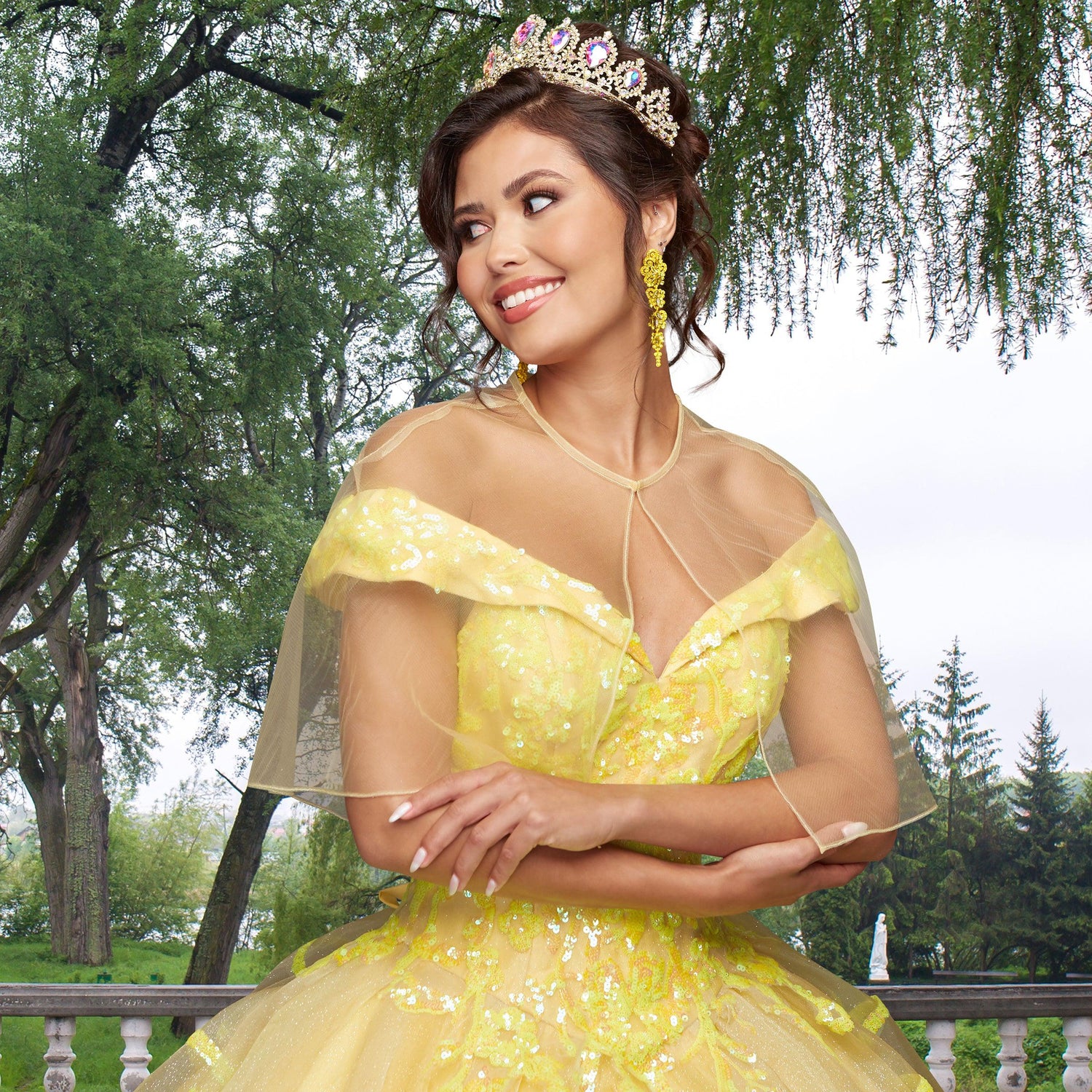 Wearing the Quinceañera Sample Dress QF104 and a jeweled tiara, a woman stands outdoors, smiling and looking to the side with trees and greenery in the background.