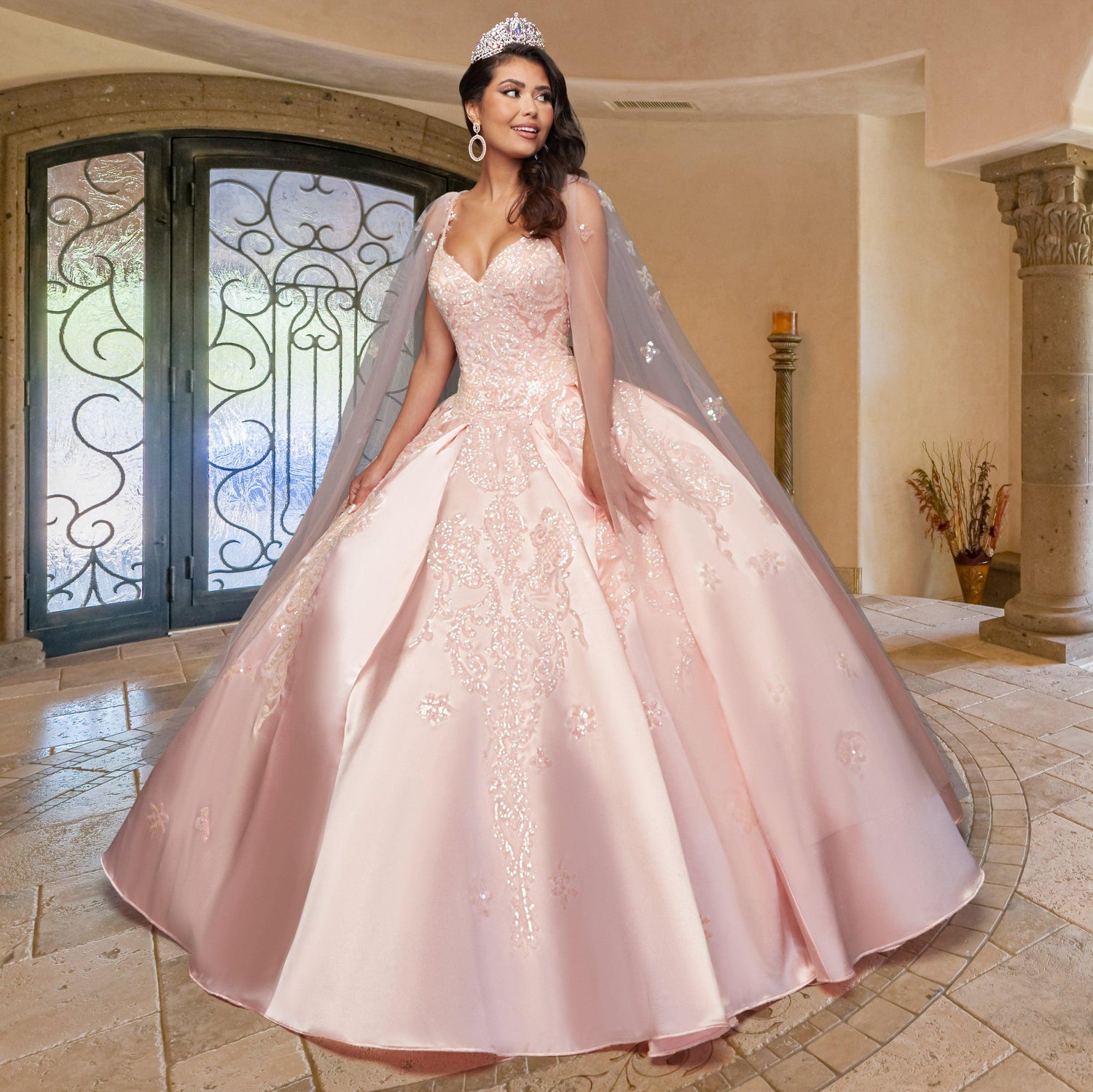 A woman wearing the Quinceañera Sample Dress QF106, a pink v-neck gown with lace details and a tiara, stands on a tiled floor indoors, smiling slightly and gazing to the side.