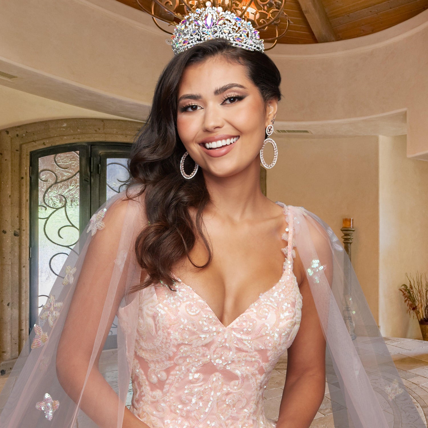 A woman indoors smiles in the Quinceañera Sample Dress QF106, a beaded light pink ball gown. She wears a jeweled crown, large earrings, and has long, dark wavy hair draped over one shoulder.