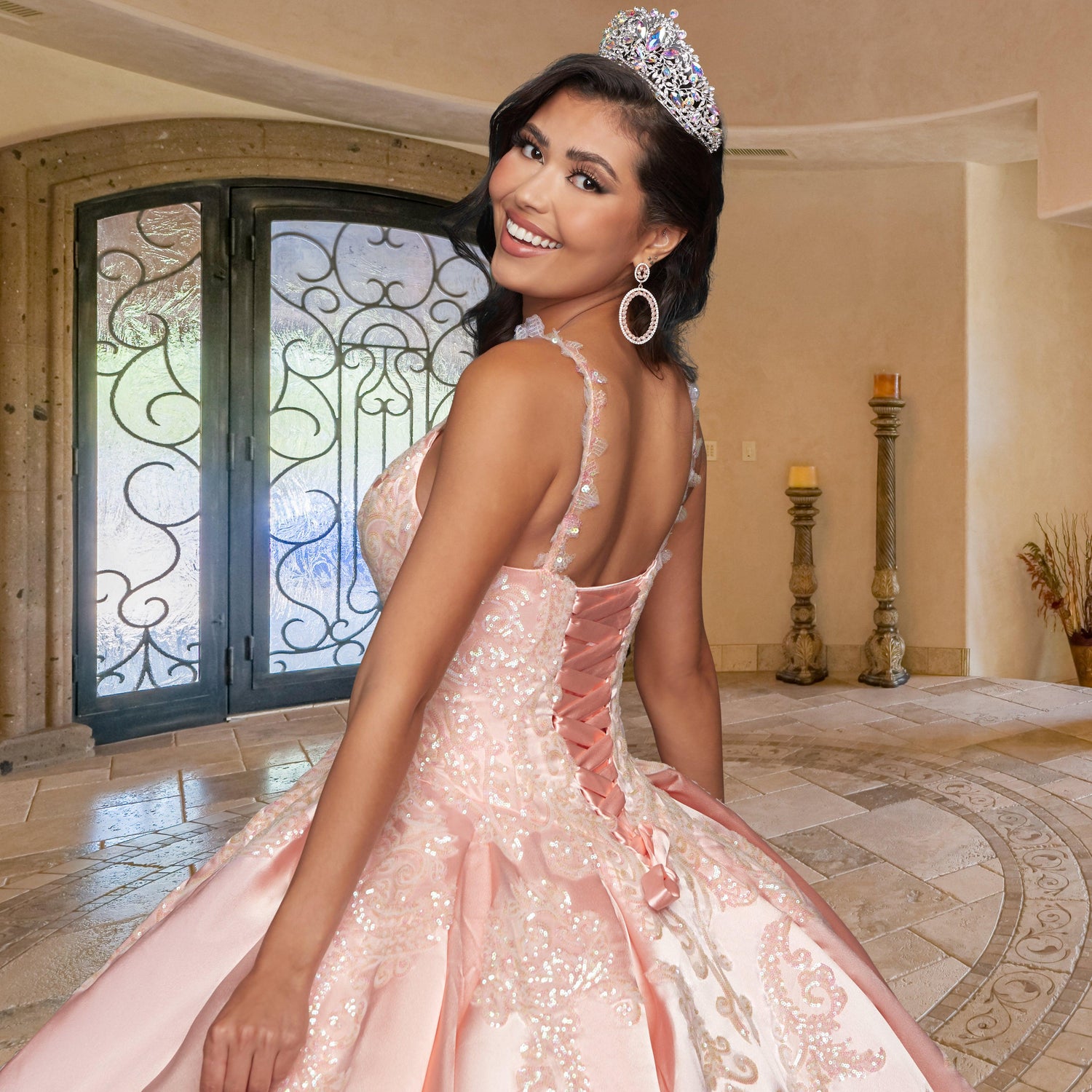 A young woman in the Quinceañera Sample Dress QF106 and a tiara smiles over her shoulder, standing in an elegantly decorated room with stone floors and ornate double doors.
