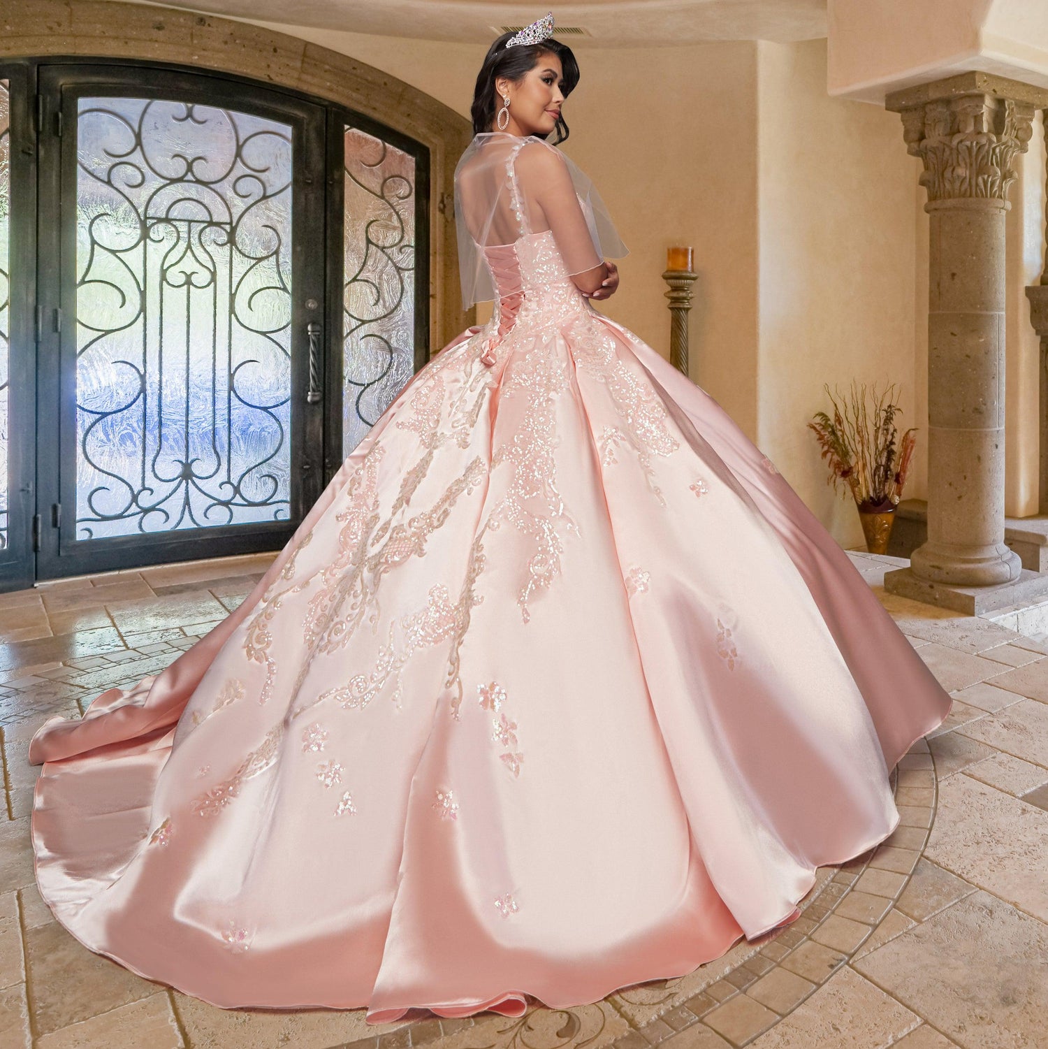 A woman wears the Quinceañera Sample Dress QF106, a pink sequin gown with embroidered details, as she stands indoors near ornate double doors and stone columns, glancing over her shoulder.