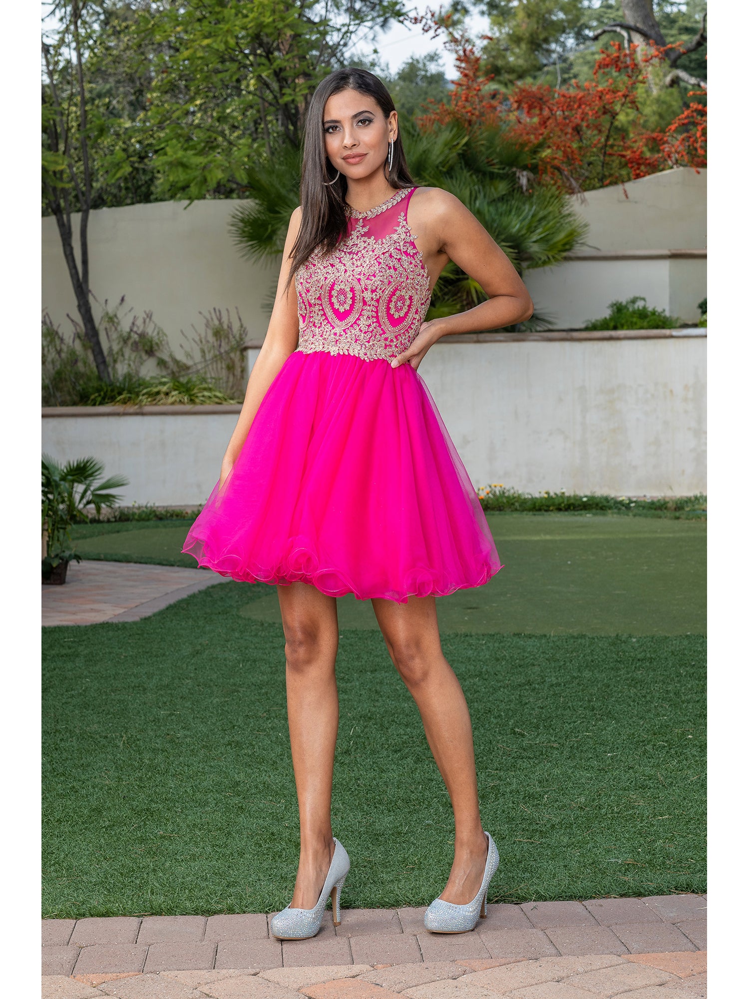 Outdoors on grass and pavement, a woman wears the Beaded Embroidered Lace Short Babydoll Party Dress in pink with silver heels. Trees and shrubbery in the background add to her elegant homecoming style.