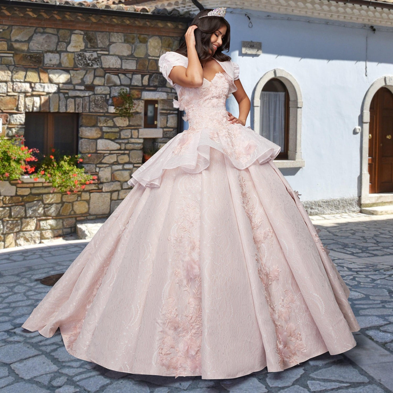 A woman wearing the Quinceañera Sample Dress QF115 and a tiara poses on a stone-paved street, set against rustic buildings with stone and plaster walls.