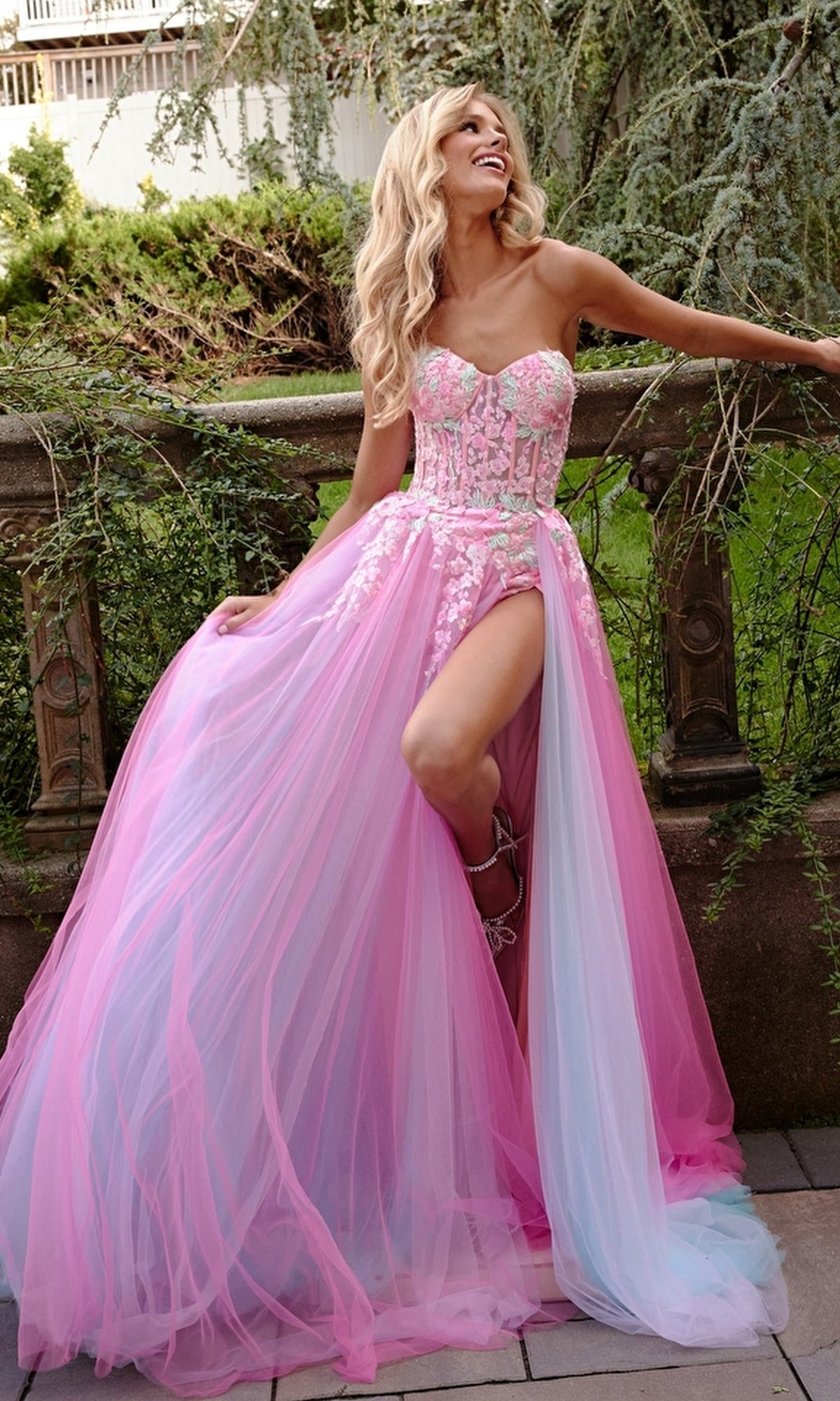 A woman in the Jovani Sheer-Corset Prom Dress with floral embroidery, featuring pink and blue tulle and a high slit, smiles as she leans against an outdoor stone railing.
