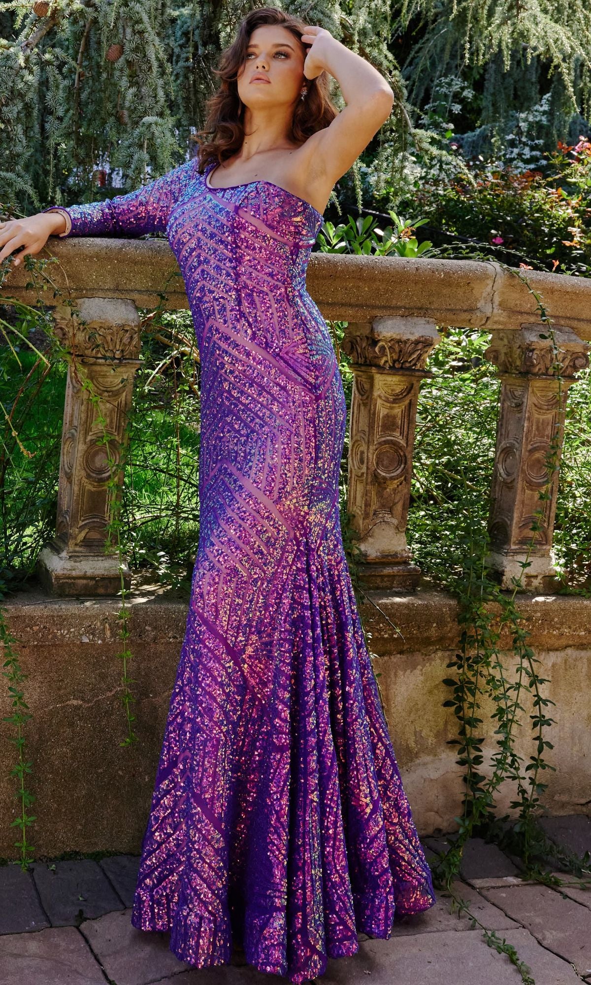 A woman wearing the Jovani Violet Purple Prom Dress with one long sleeve, accented with sequins, stands outdoors by a stone balustrade surrounded by greenery and flowers.