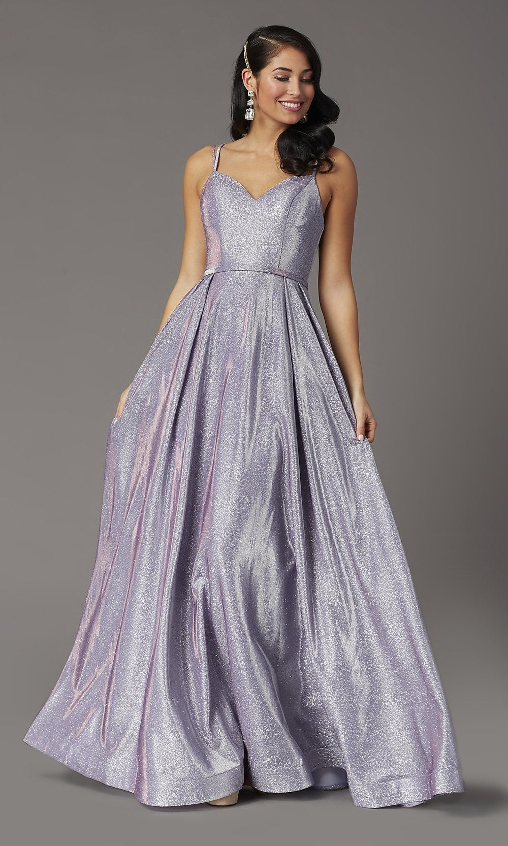 A woman poses against a plain gray background in a Metallic Long A-Line Prom Dress, featuring a floor-length shimmery lavender jersey, spaghetti straps, fitted bodice, and side pockets.