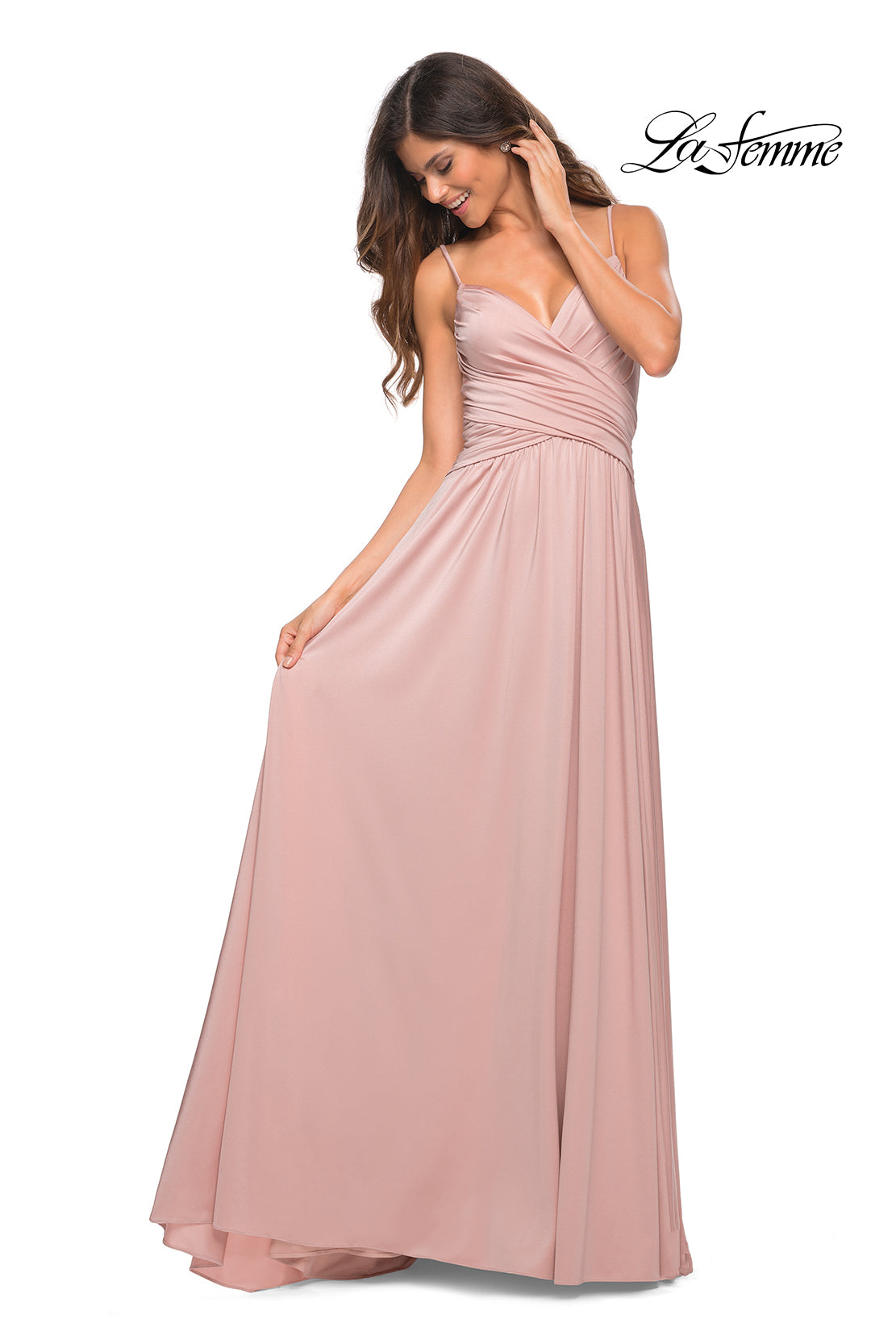 A woman models the La Femme Simple Elegant Long Formal Prom Dress in blush pink, featuring a floor-length, sleeveless design with a ruched bodice and v-neckline. She poses against a white background, gracefully holding one side of the skirt.