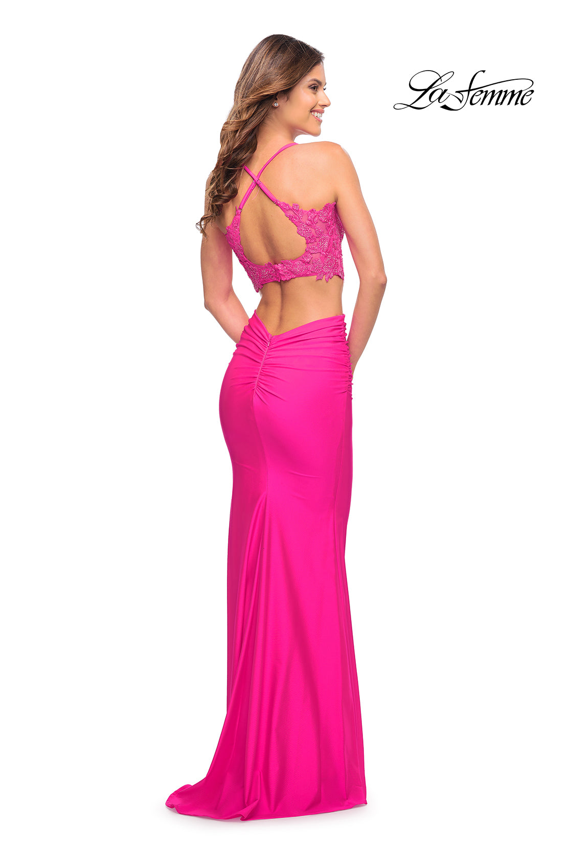 A woman stands looking over her shoulder in a Neon Pink Two-Piece Long La Femme Prom Dress with a lace crop top and open back. The