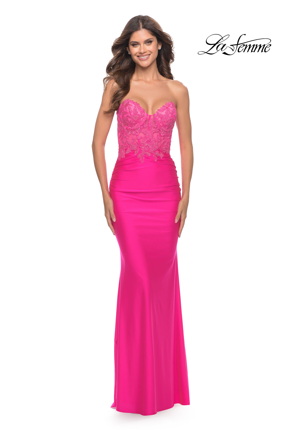 A woman smiles against a white background wearing the La Femme Strapless Neon Pink Prom Dress with lace detailing on the bodice. The La Femme logo appears in the top right corner.