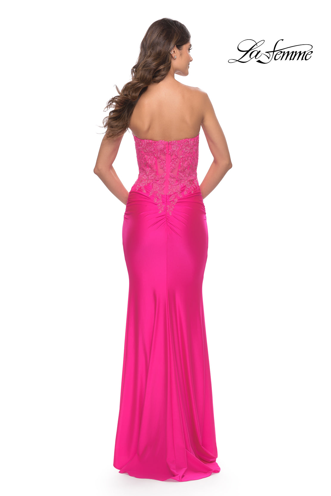 A woman stands with her back to the camera, wearing the La Femme Strapless Neon Pink Prom Dress featuring lace detailing on the bodice.