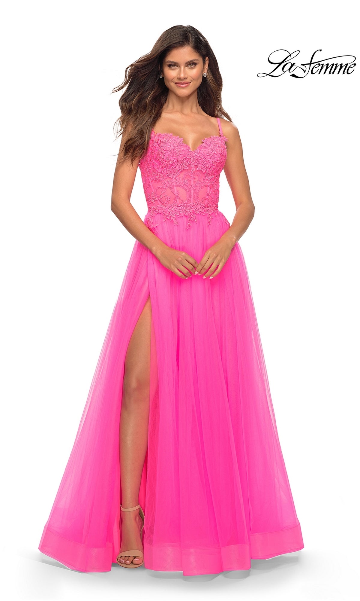 A woman models the La Femme Neon Pink Long Ball Gown with Lace, featuring a sheer bodice, lace details, and a high slit, posing against a white backdrop with the La Femme logo for a striking prom dress look.