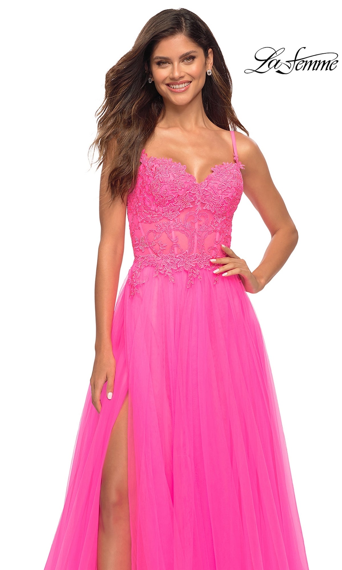A woman smiles confidently in the La Femme Neon Pink Long Ball Gown with Lace, featuring a slit skirt and lace bodice. The La Femme logo appears in the top right corner.