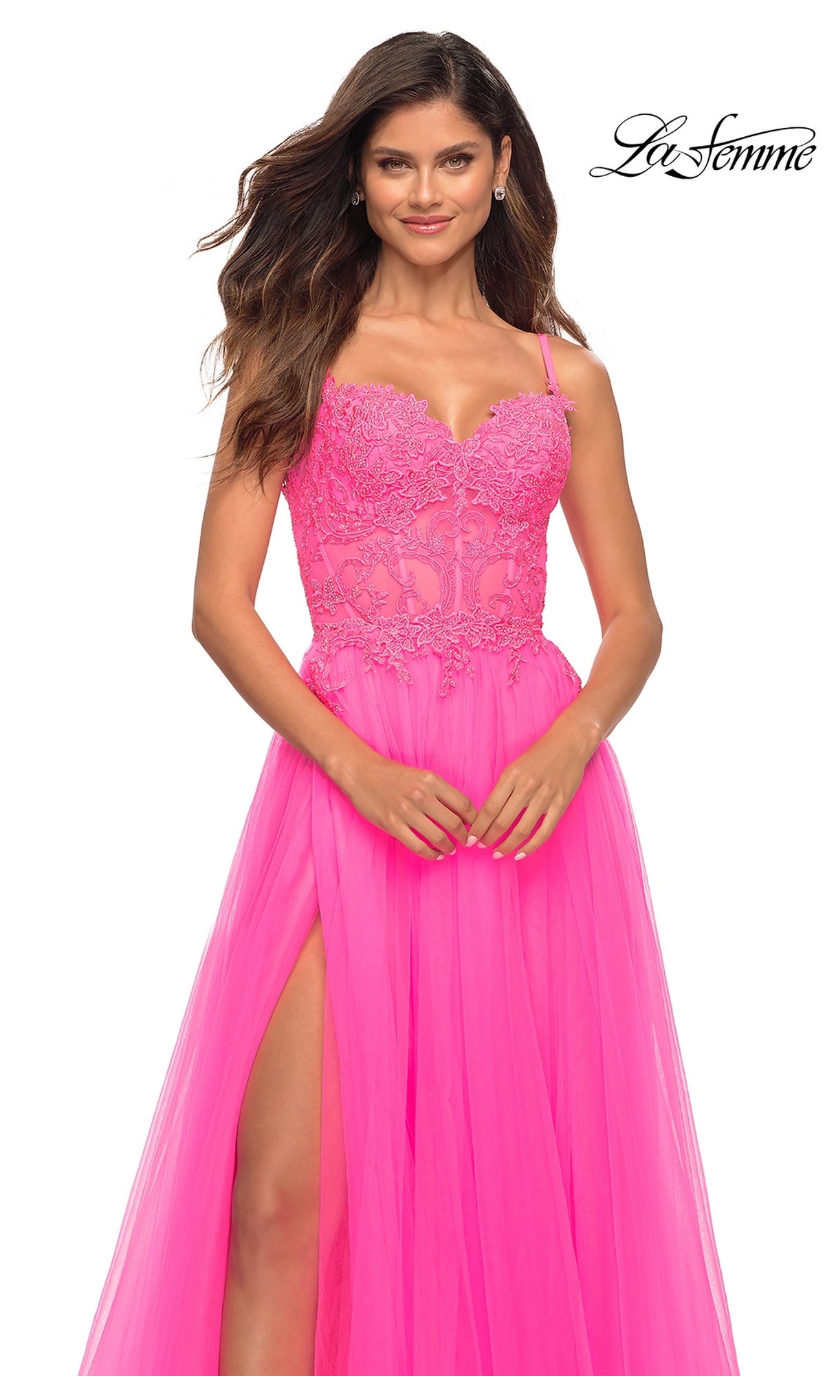 A woman models the La Femme Neon Pink Long Ball Gown with Lace, featuring spaghetti straps, a lace bodice, and a high slit, set against a white background. The