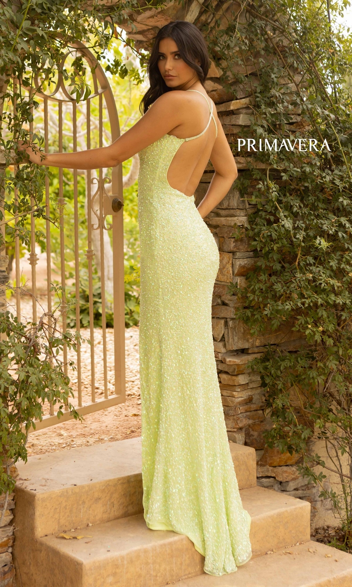 A woman stands outdoors by a stone arch and iron gate, surrounded by lush greenery, wearing the Deep V-Neck Long Sequin Prom Dress with Slit in fitted light green with thin straps.