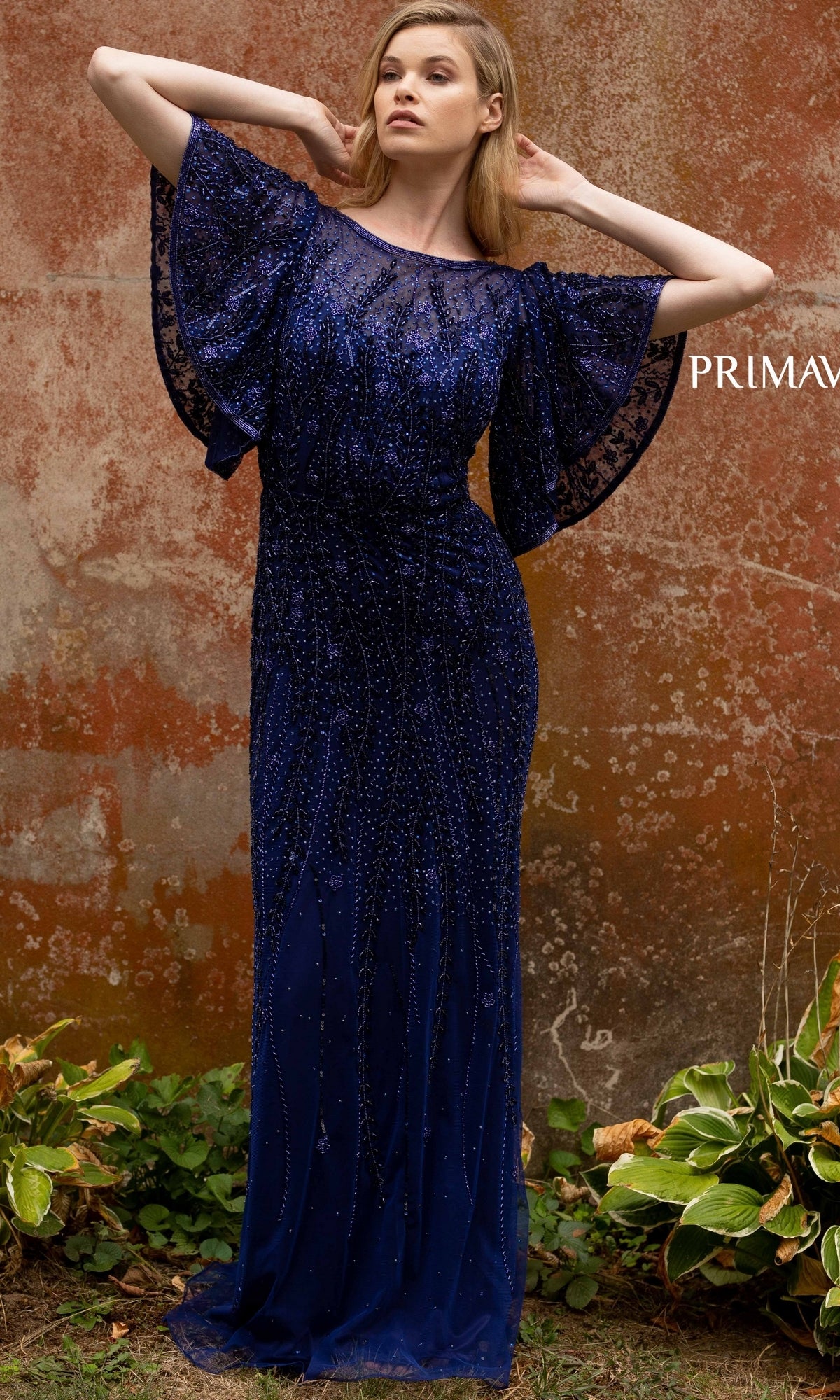 A woman stands outdoors by a weathered wall, wearing the elegant Short-Sleeve Beaded Long Prom Dress 3681. This floor-length navy gown features flutter sleeves and delicate detailing, with green plants at her feet.