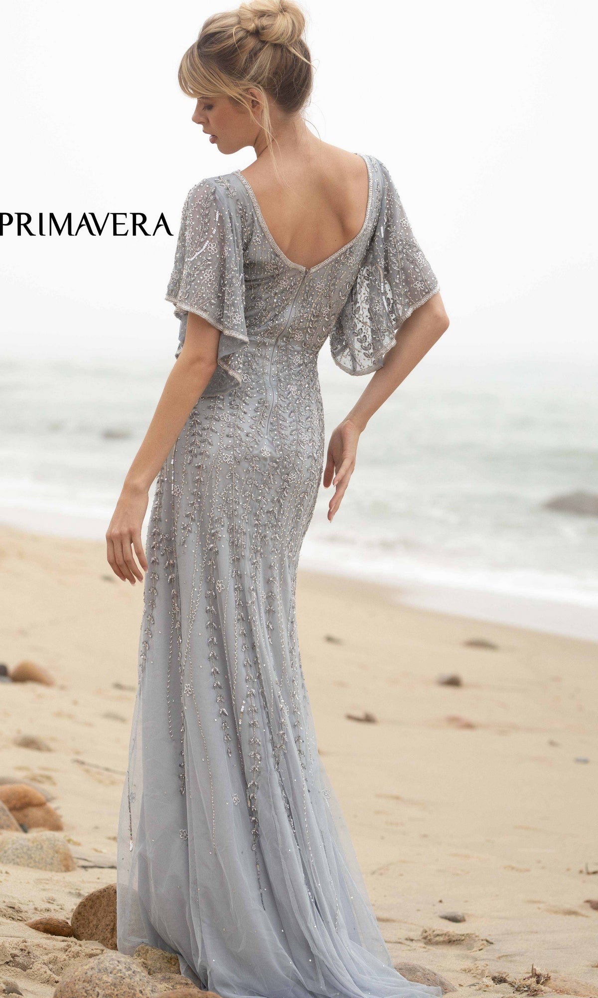 A woman stands on a sandy beach facing the ocean, wearing the Short-Sleeve Beaded Long Prom Dress 3681, its silver beading shimmering in the light as her hair is styled in an elegant updo.