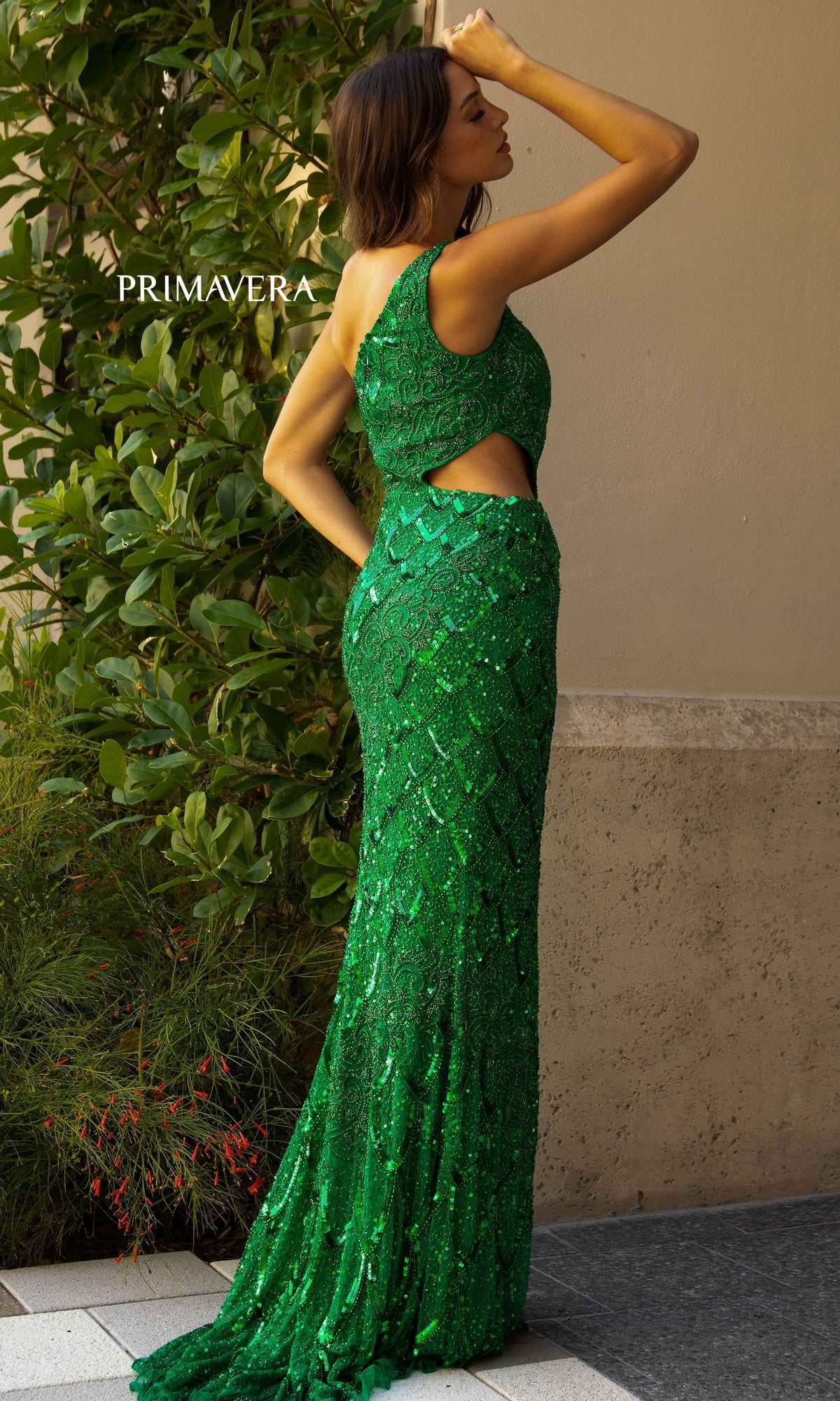 A woman stands outdoors near a wall and greenery, wearing the Primavera 3729 One-Shoulder Long Sequin Prom Dress in green with a side cutout.
