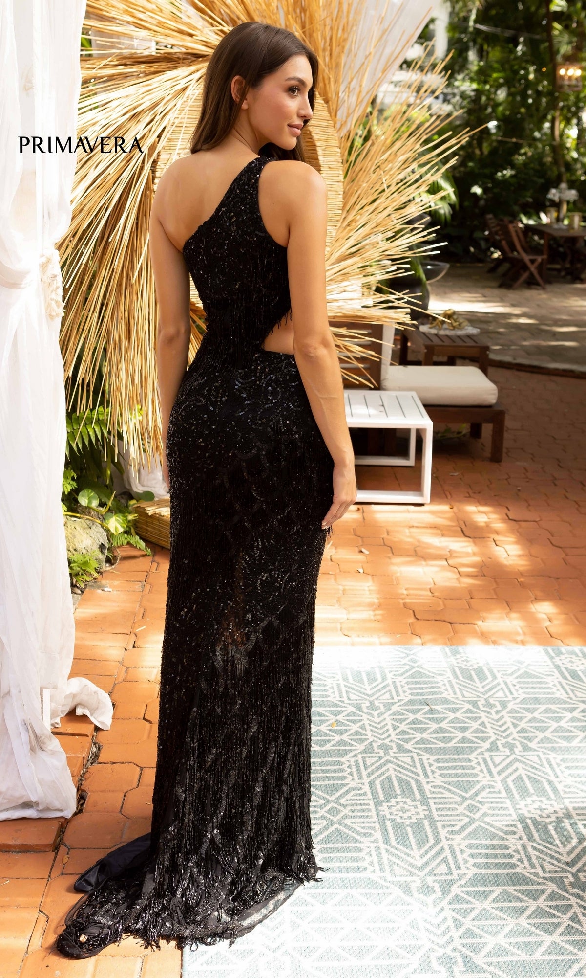 A woman stands on a brick patio, facing away in the One-Shoulder Long Beaded Prom Dress with Fringe. This dazzling black designer gown features floor-length sequins for an elegant outdoor look.