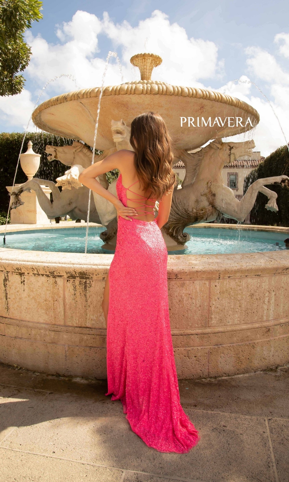 A woman in the Bright Wrap-Style Long Sequin Prom Dress 3791B stands with her back to the camera in front of a decorative fountain on a sunny day.