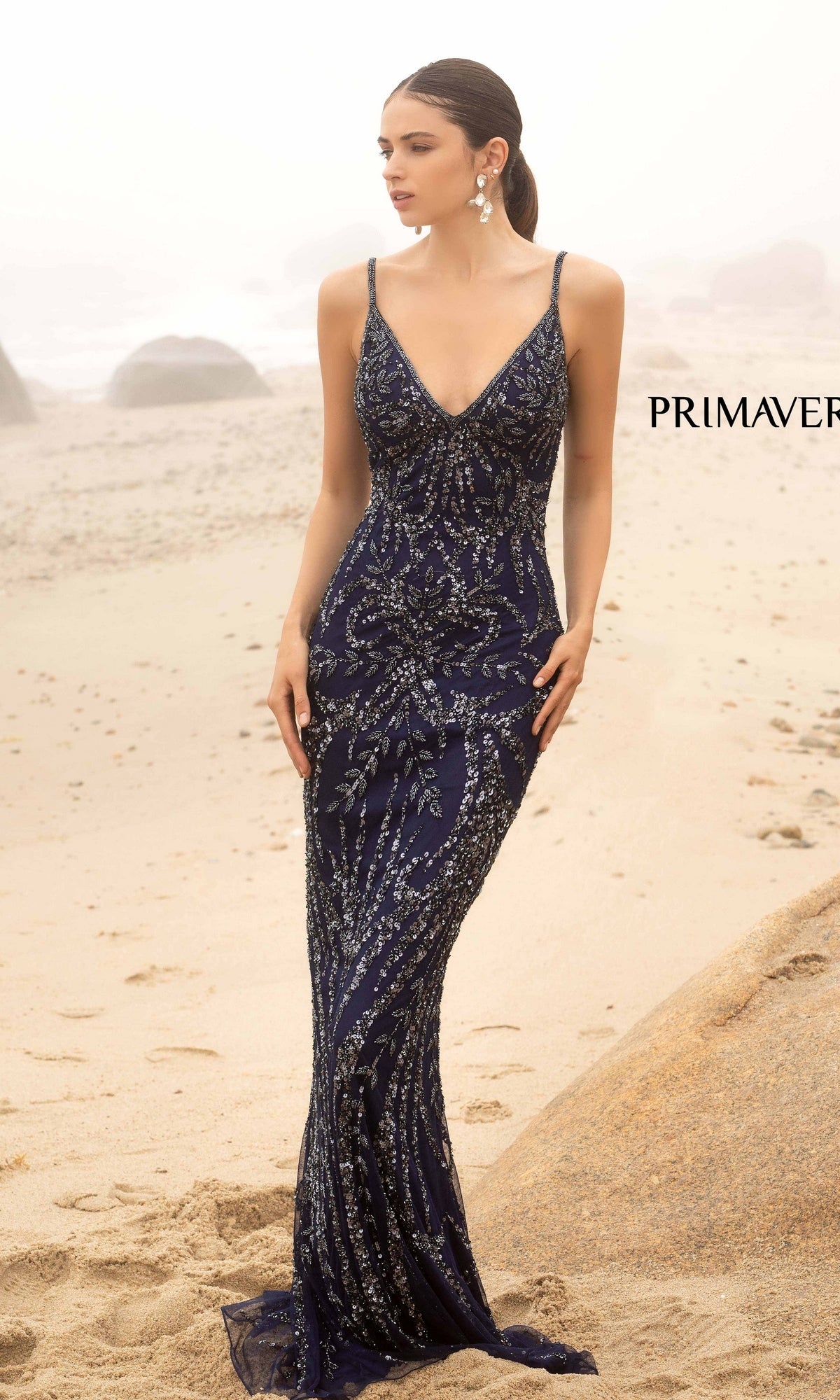 A woman wears the Primavera V-Neck Long Sequin Prom Dress 3793D, standing on a sandy beach with rocks in the background.