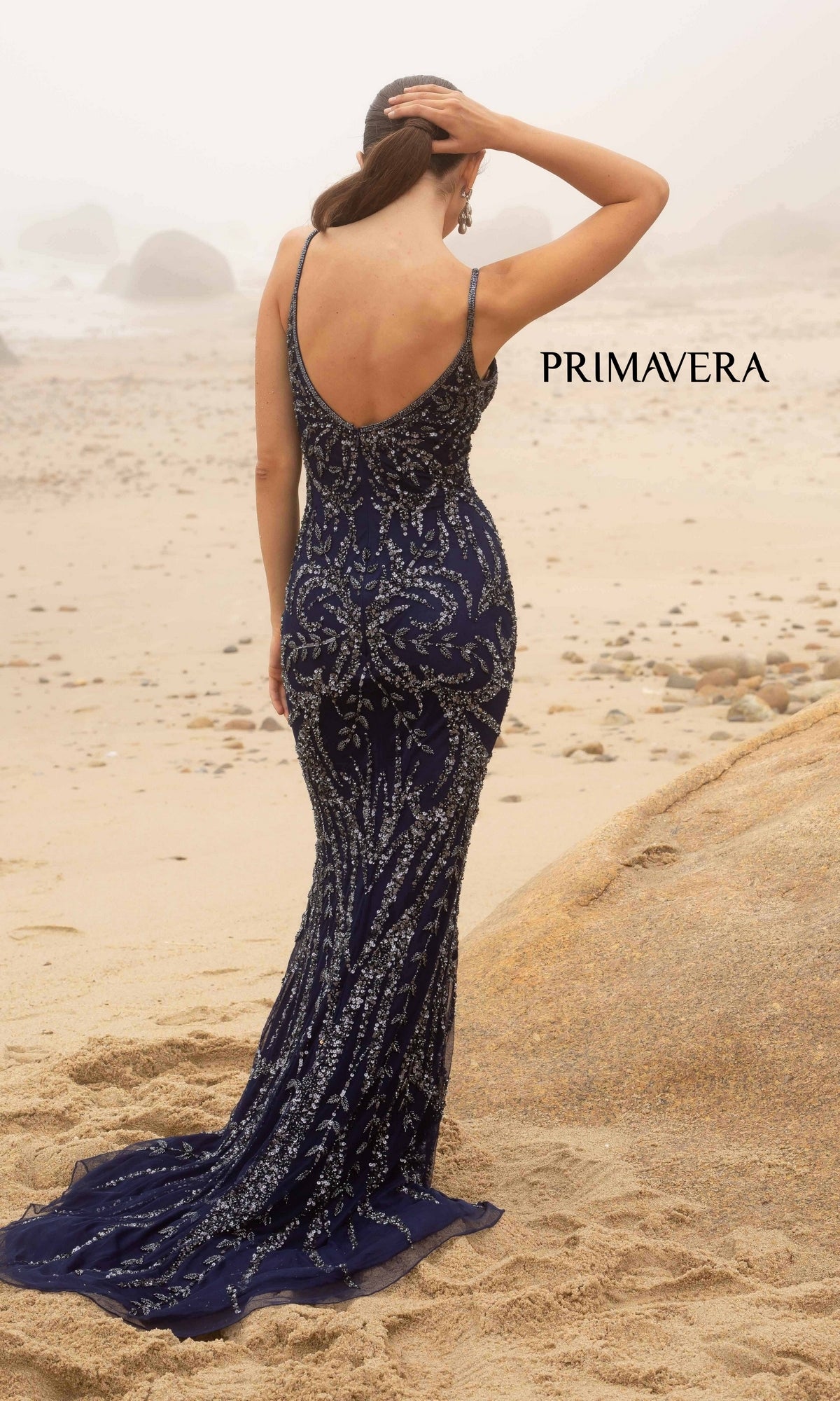 A woman in a navy blue Primavera V-Neck Long Sequin Prom Dress 3793D stands on a sandy beach facing away, hand on her head.