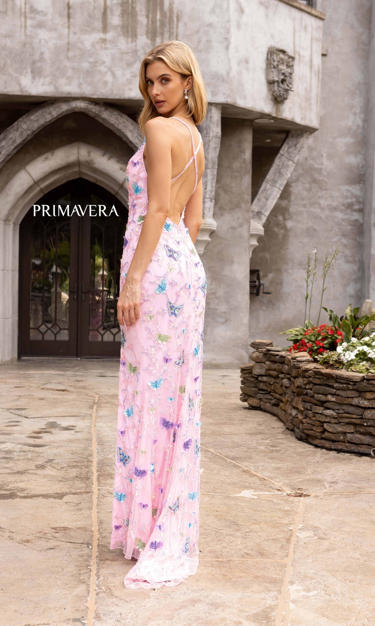 A woman in the Beaded Butterfly Long Prom Dress by Primavera 3901, featuring spaghetti straps and a flowing skirt with a delicate butterfly pattern, stands outside a stone building, glancing over her shoulder.